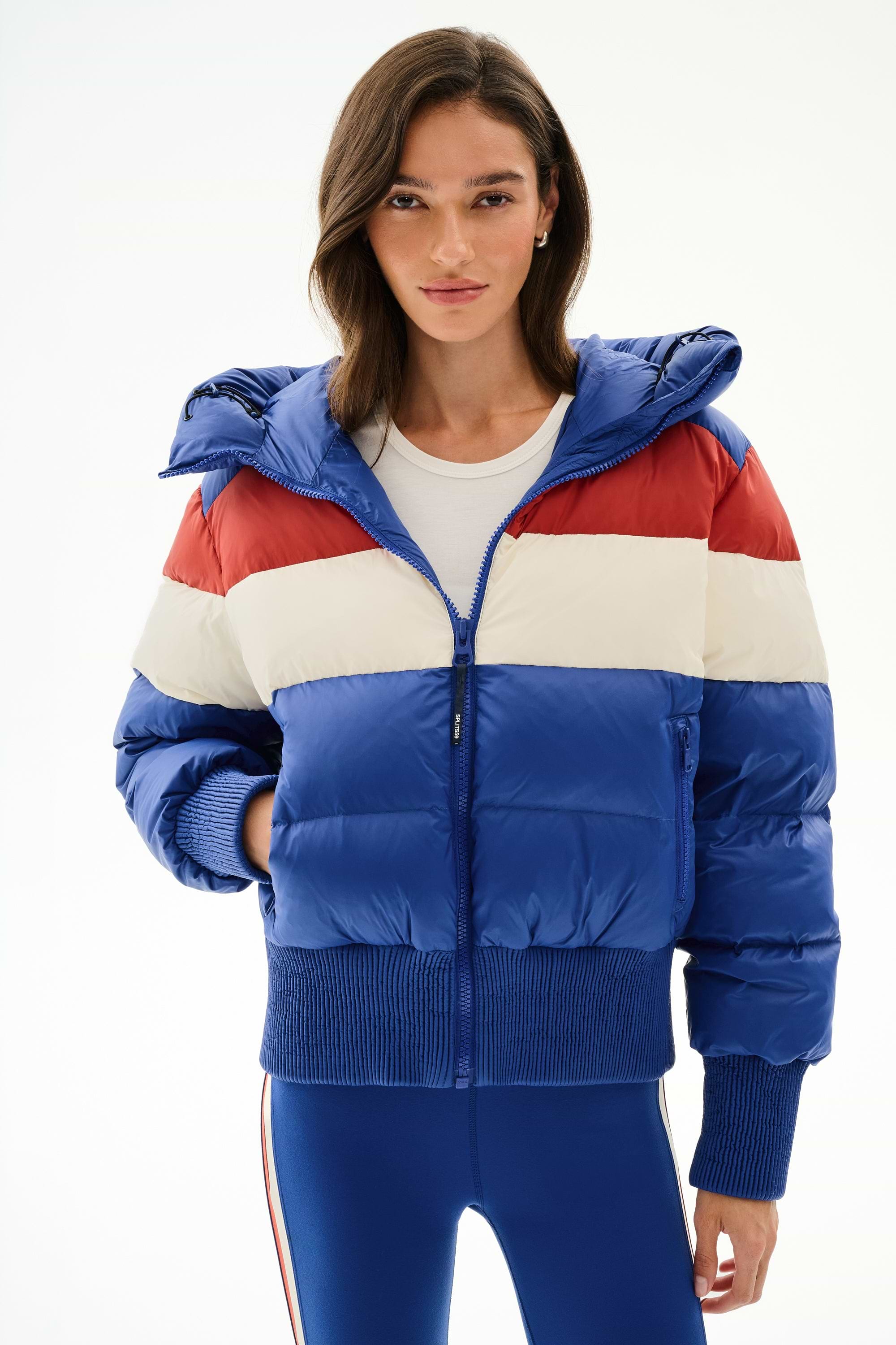 A woman in the SPLITS59 Aspen Puffer Jacket - Cobalt/Creme, a nylon, white duck down jacket, with a white shirt and blue leggings against a plain white background.