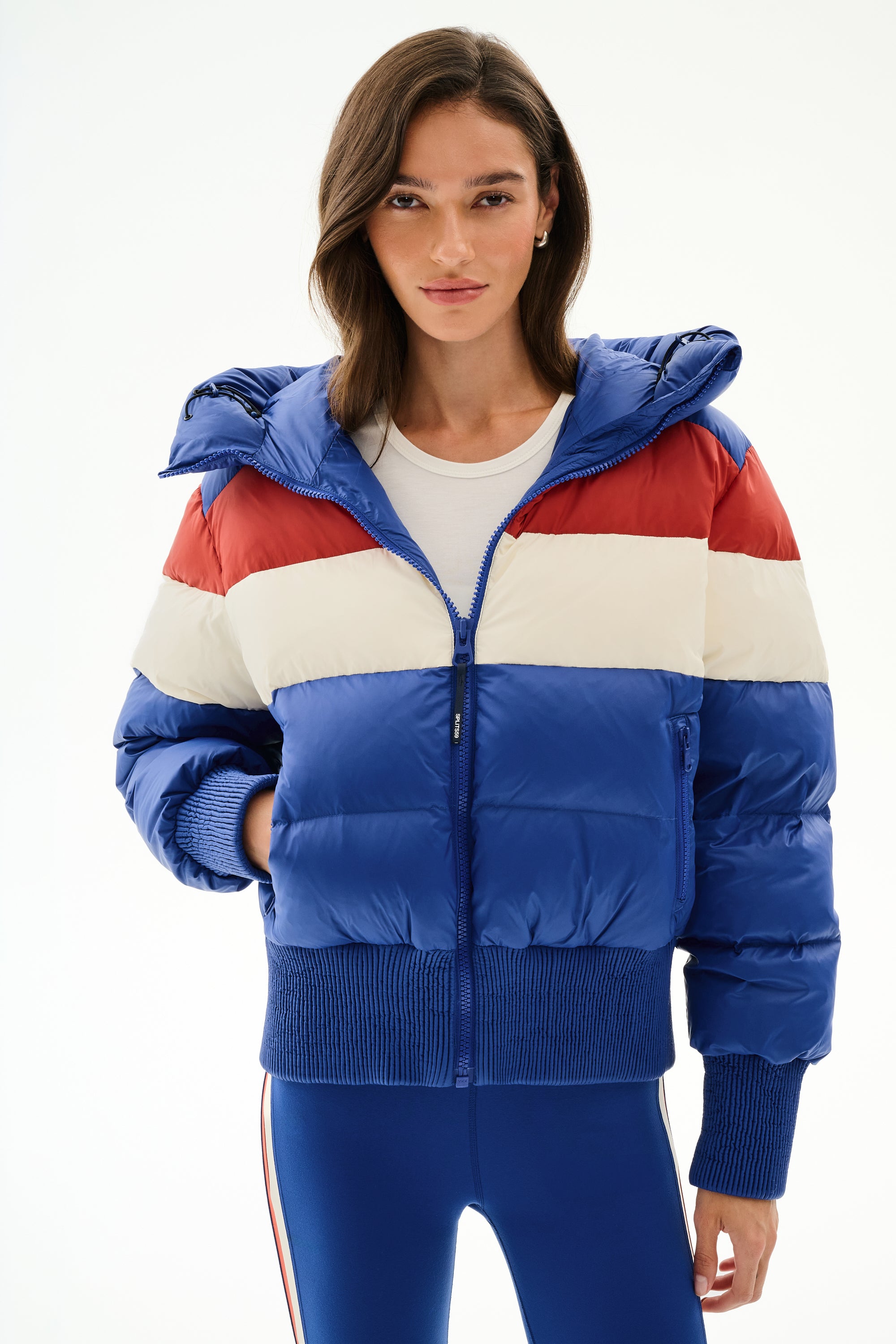A woman in the SPLITS59 Aspen Puffer Jacket - Cobalt/Creme, a nylon, white duck down jacket, with a white shirt and blue leggings against a plain white background.