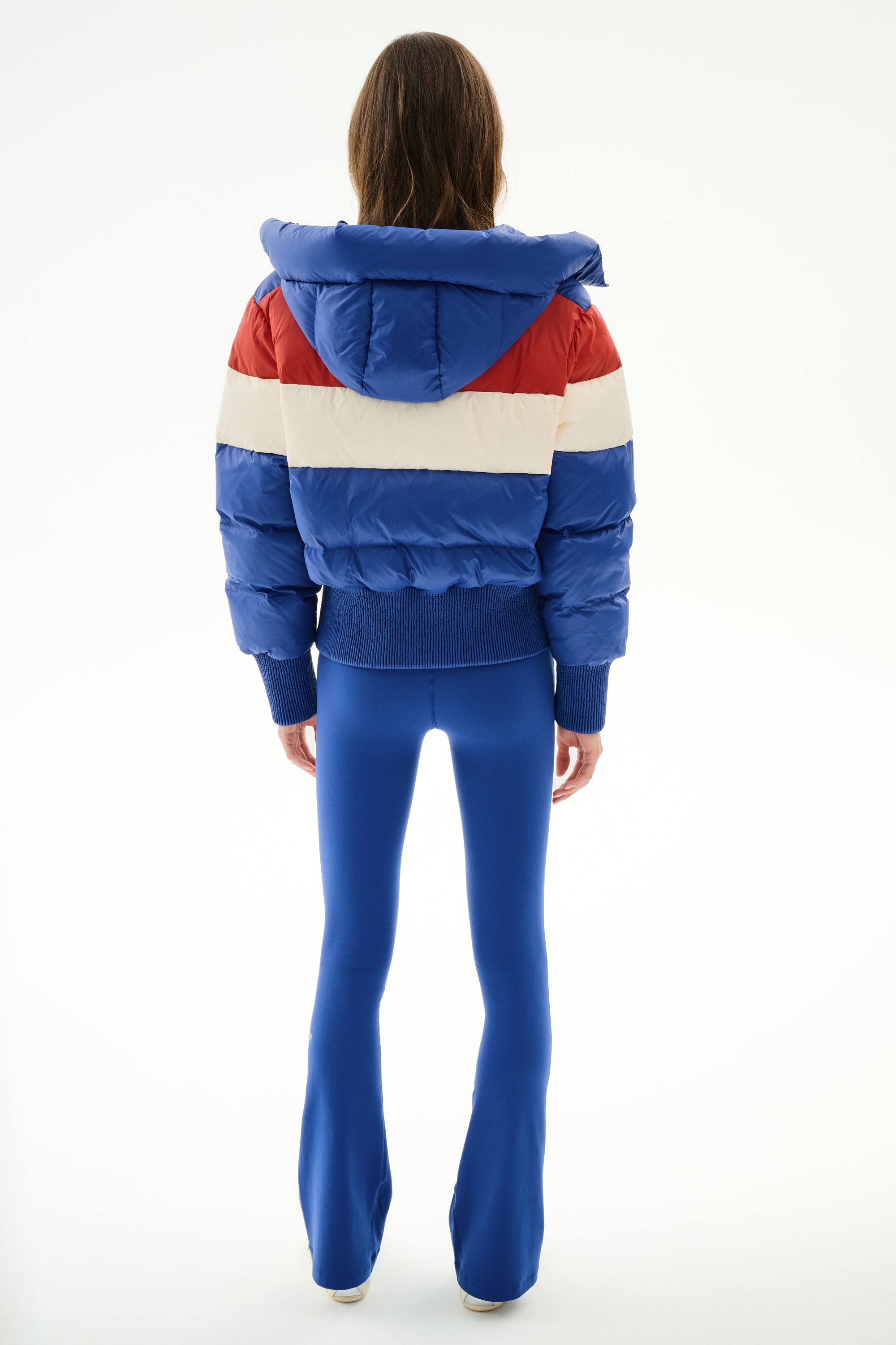 Person facing away, wearing the SPLITS59 Aspen Puffer Jacket - Cobalt/Creme with a hood and bright blue flared pants, standing against a plain white background.