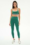 A woman stands against a white background, wearing the SPLITS59 Bianca Rigor Bra in Arugula/White with matching leggings in a soft nylon spandex blend, paired with white sneakers.
