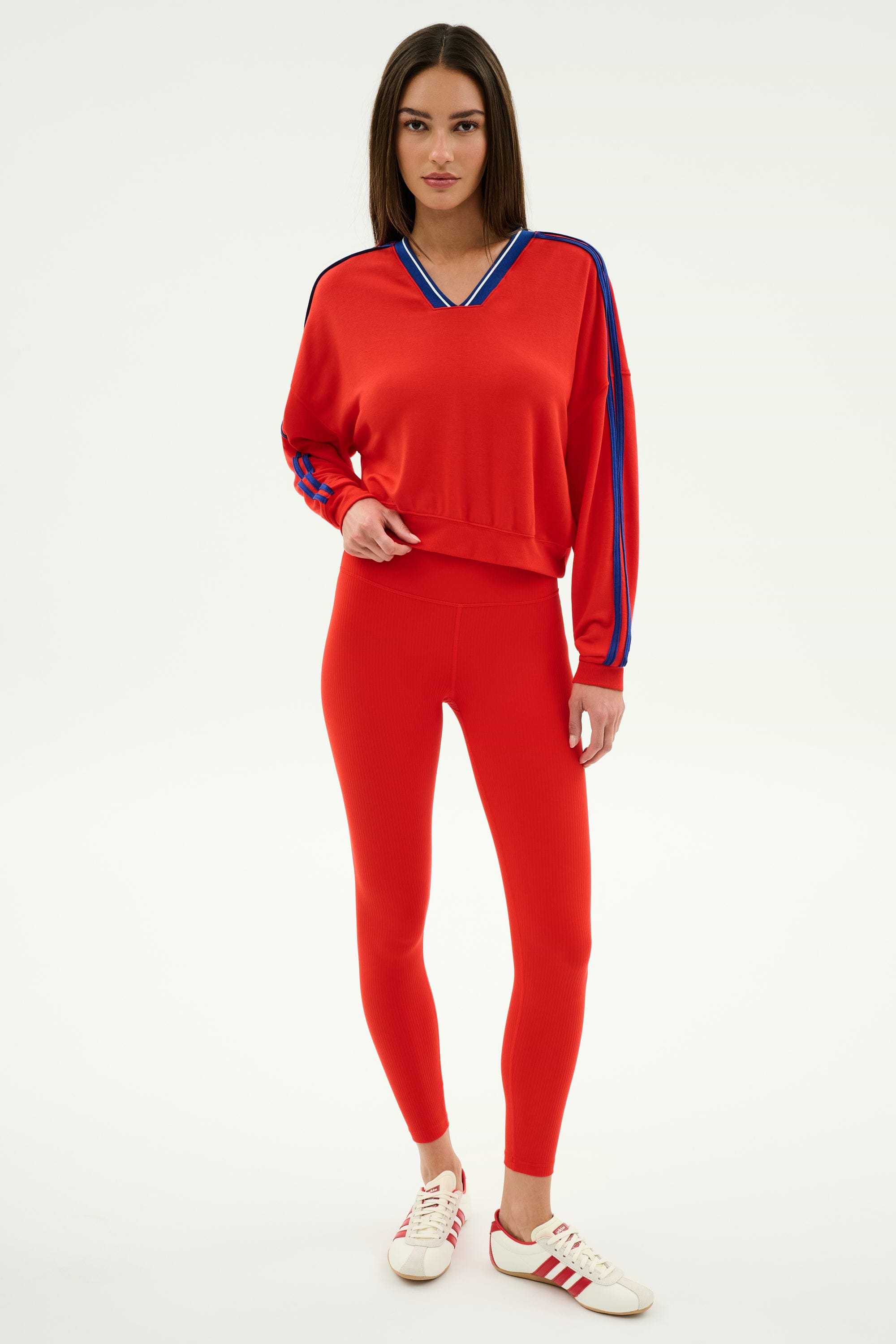 A woman wears the SPLITS59 Ellie Fleece V Neck Sweatshirt in Lotus Red/Cobalt, paired with red leggings and white sneakers with red stripes, standing against a plain white background.