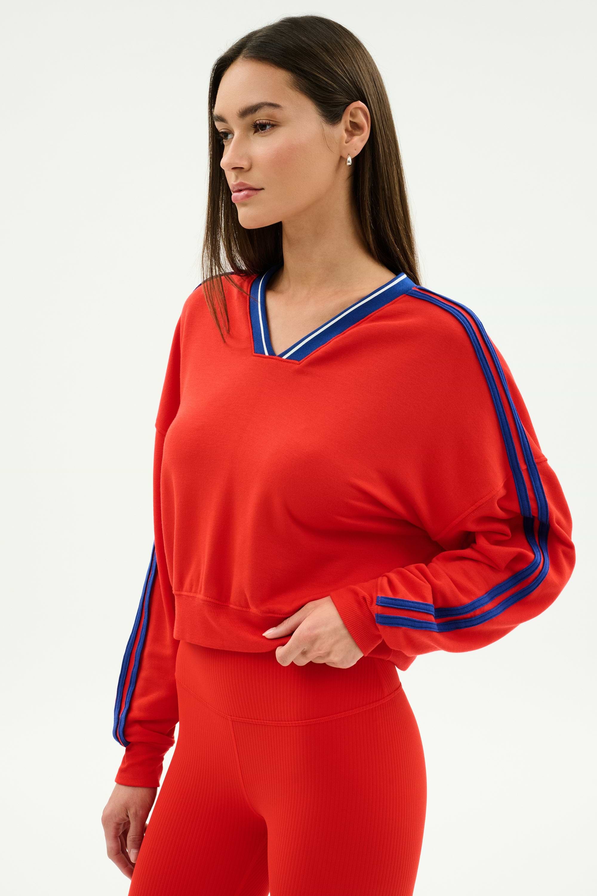 A woman wears the SPLITS59 Ellie Fleece V Neck Sweatshirt in Lotus Red/Cobalt with blue sleeve stripes and matching red spandex-blend leggings, standing against a plain light background.