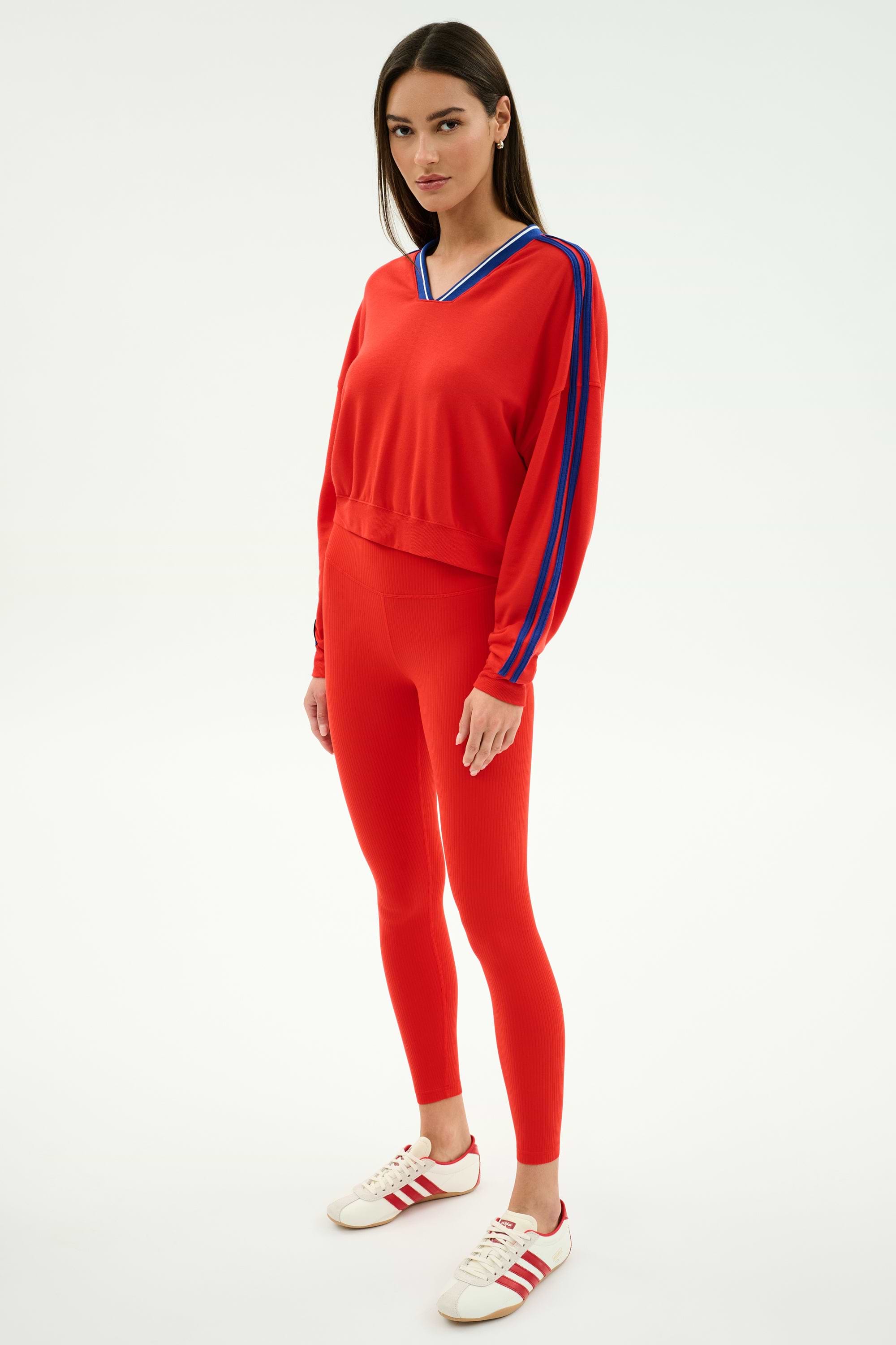 A woman wears the SPLITS59 Ellie Fleece V Neck Sweatshirt in Lotus Red/Cobalt, featuring blue sleeve stripes, paired with matching red leggings and white sneakers with red stripes.