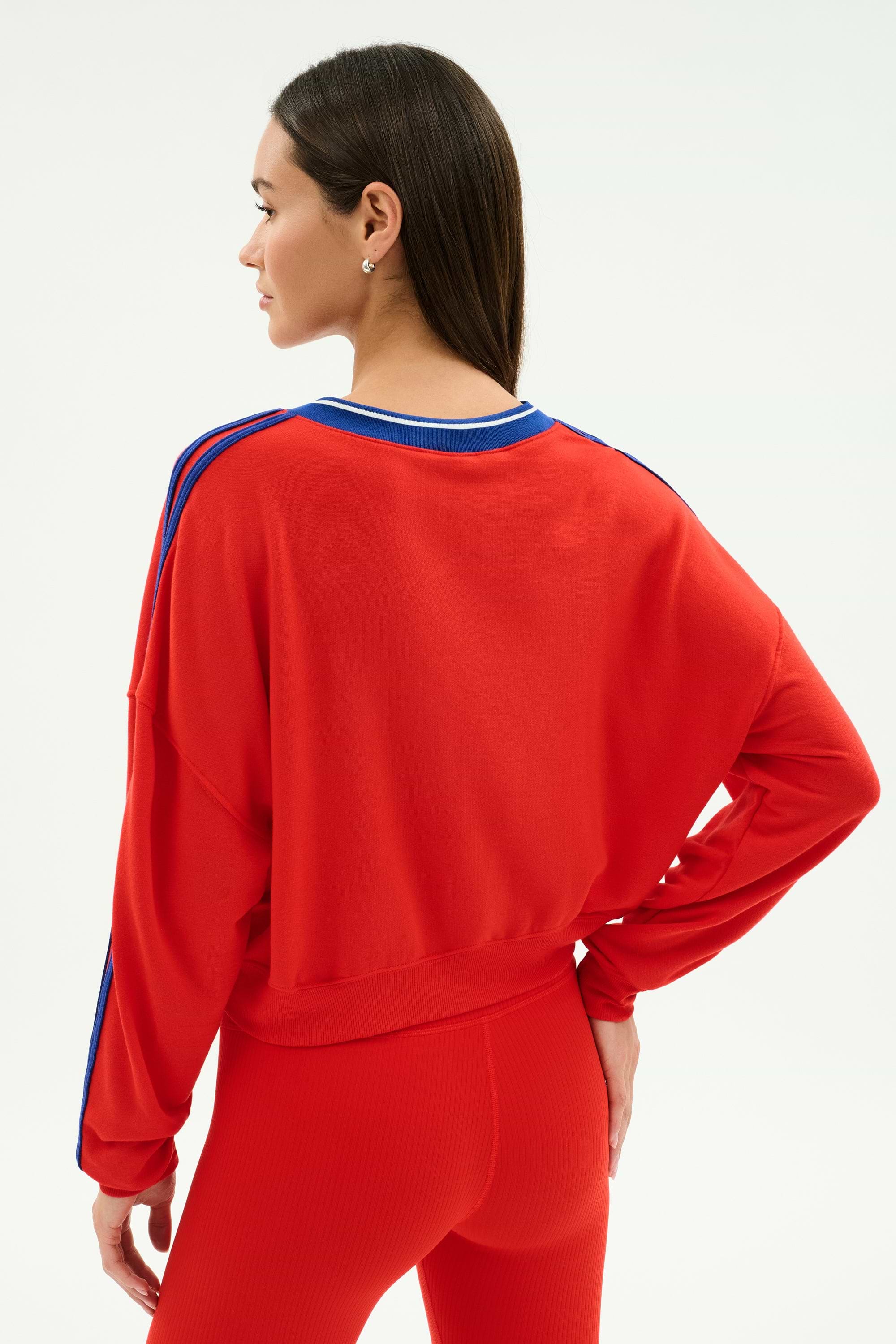 A woman stands with her back to the camera, hand on hip, wearing the SPLITS59 Ellie Fleece V Neck Sweatshirt in Lotus Red/Cobalt.