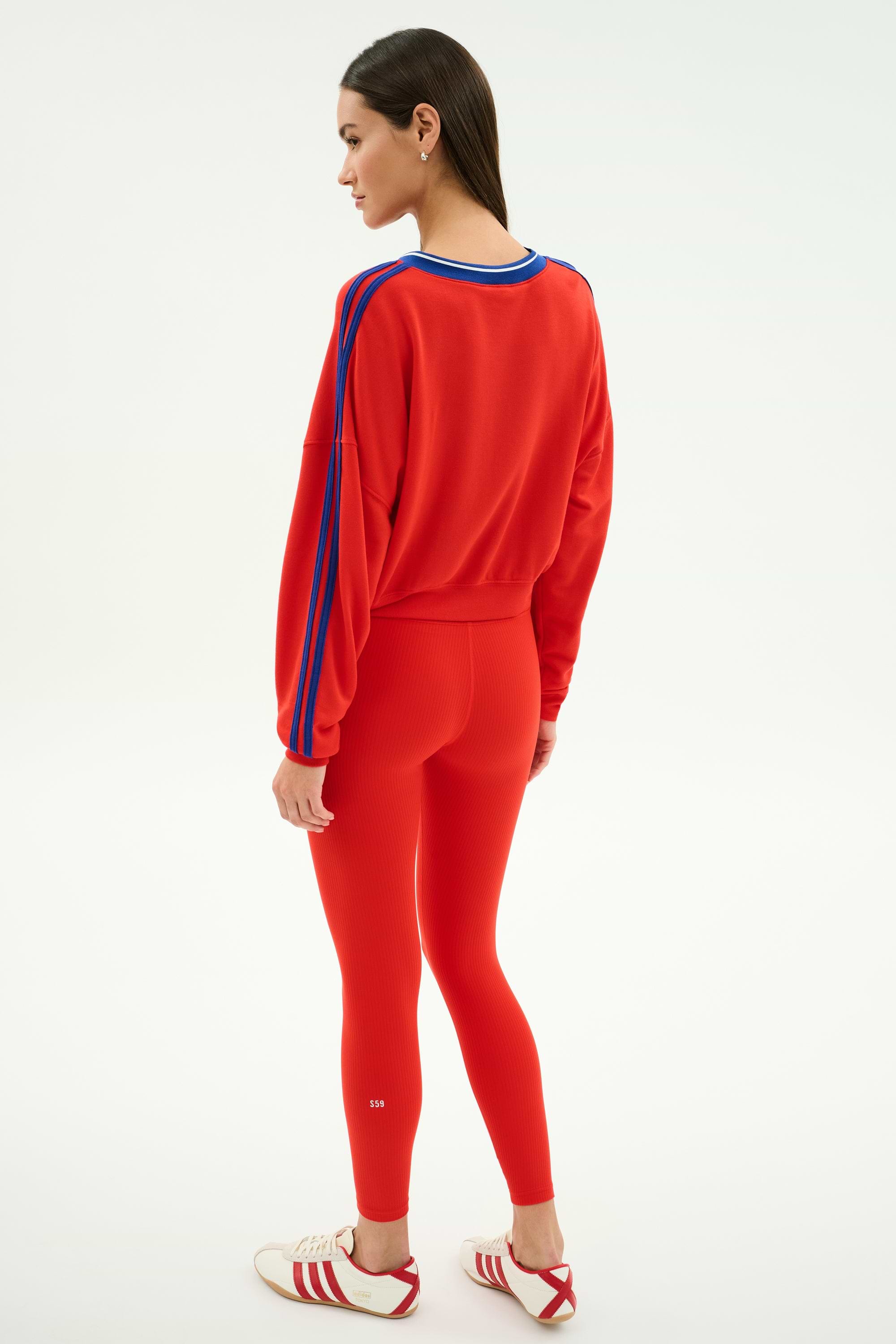 A woman stands sideways against a white background, wearing the SPLITS59 Ellie Fleece V Neck Sweatshirt in Lotus Red/Cobalt with matching red leggings and white Adidas sneakers featuring red stripes.