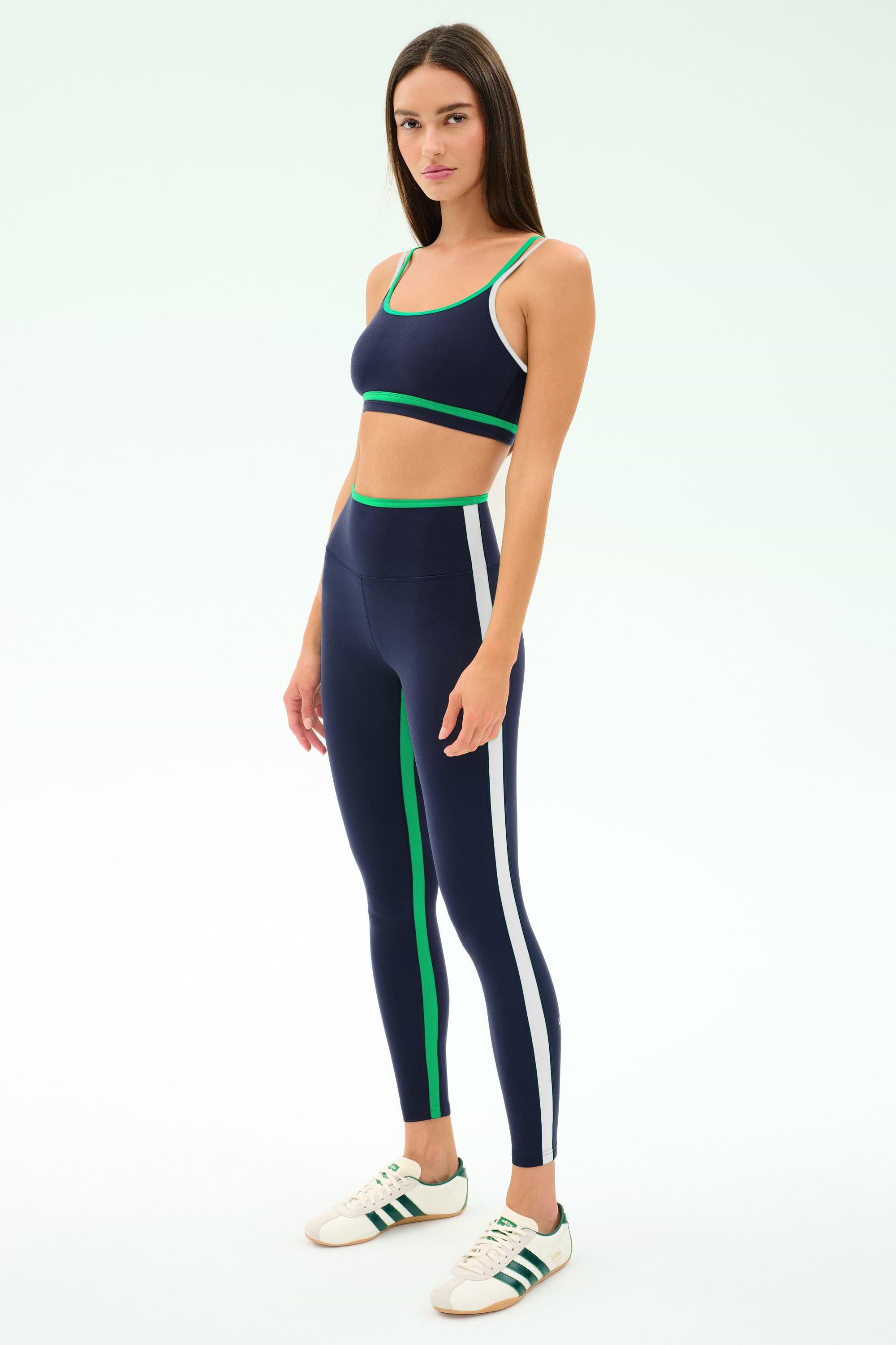 A woman models the SPLITS59 Eva Rigor Double Strap Bra in Indigo/Spearmint, paired with leggings featuring green and white stripes and white sneakers, against a plain light background.