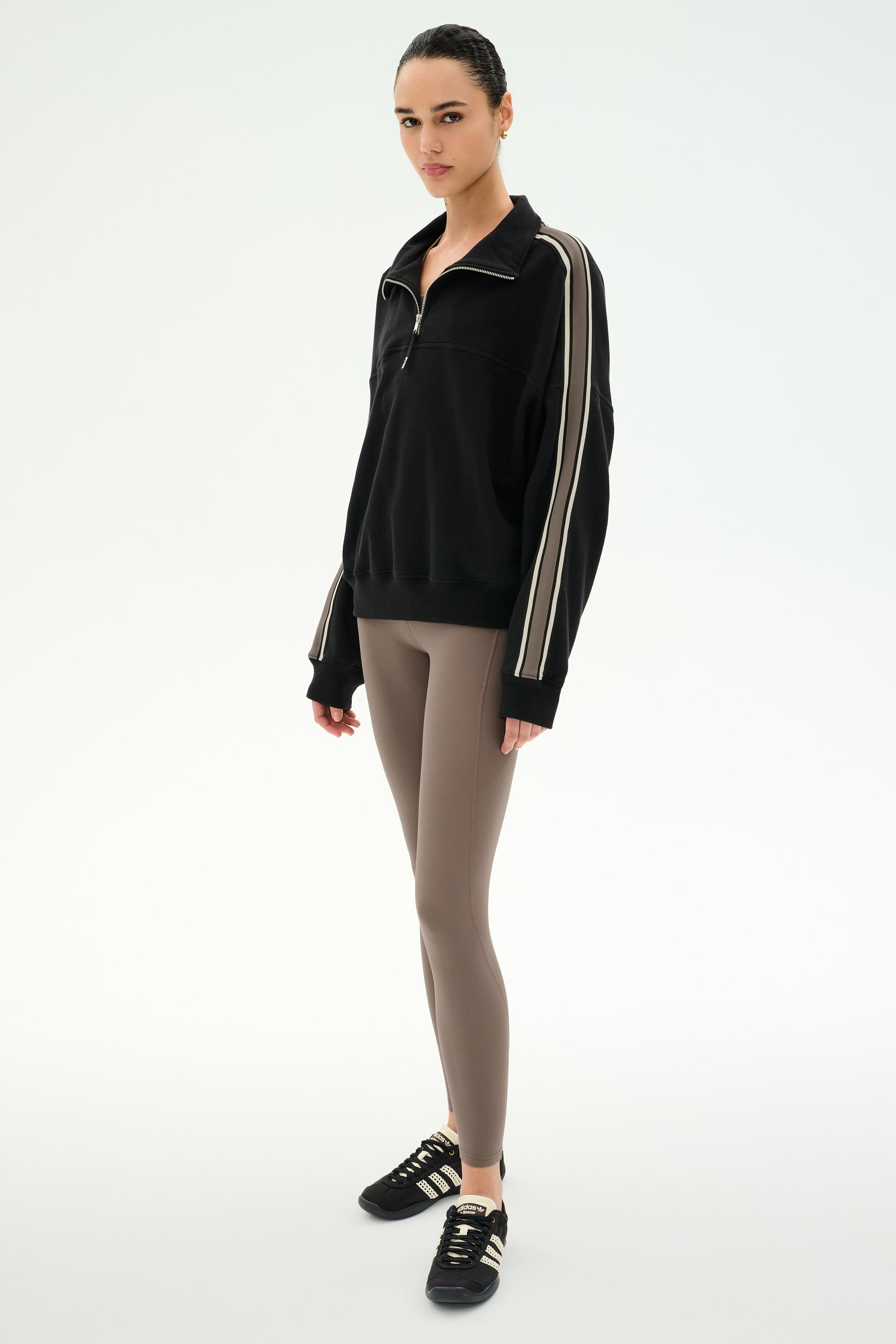 Standing against a white background, the individual is wearing the SPLITS59 Goldie French Terry Oversized Half Zip in Black/Lentil, featuring striped sleeves and soft cotton fabric, paired with taupe leggings and black Adidas sneakers.