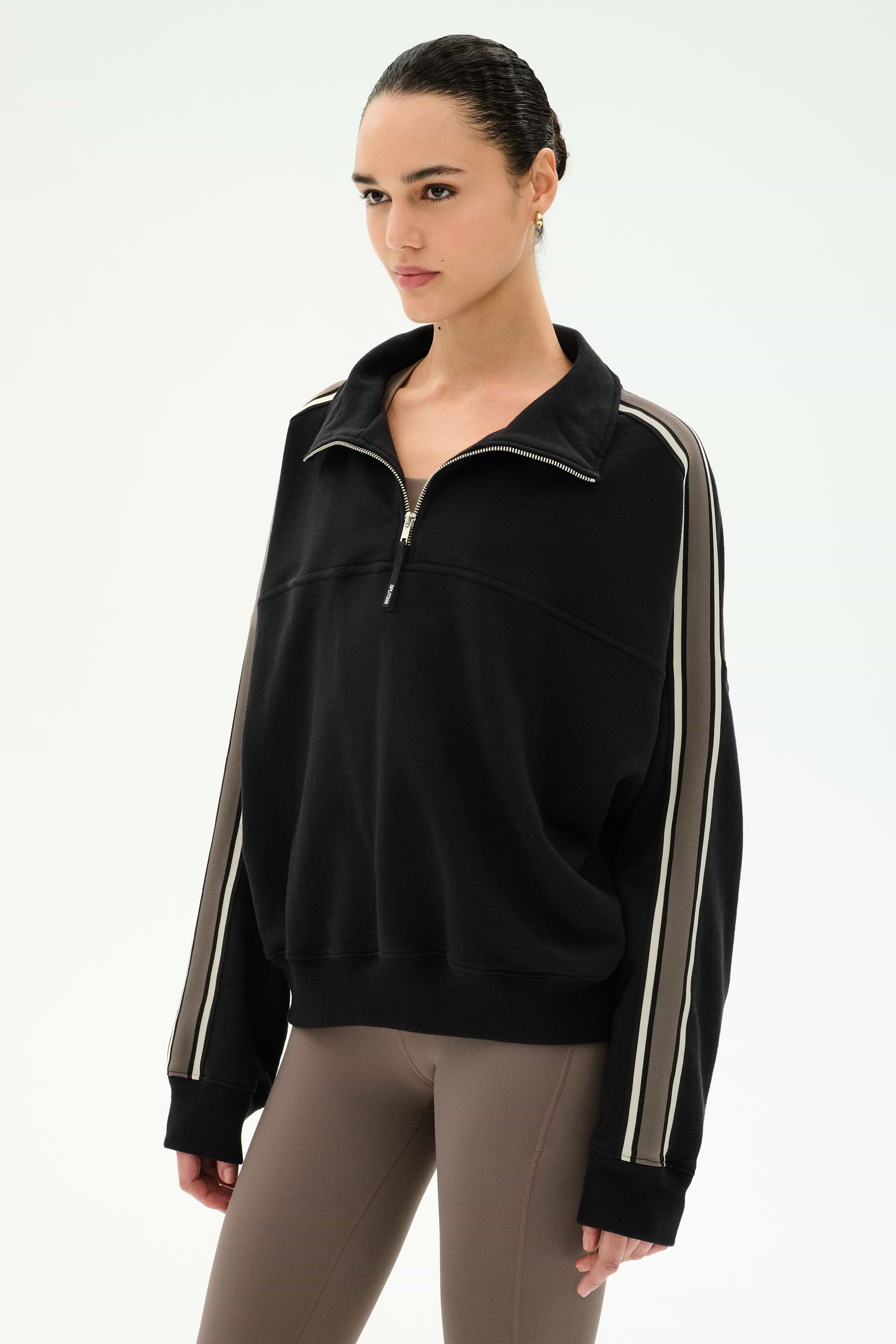 A woman wearing the SPLITS59 Goldie French Terry Oversized Half Zip in Black/Lentil, featuring soft cotton fabric and beige striped sleeves, paired with taupe leggings, stands against a plain white background.