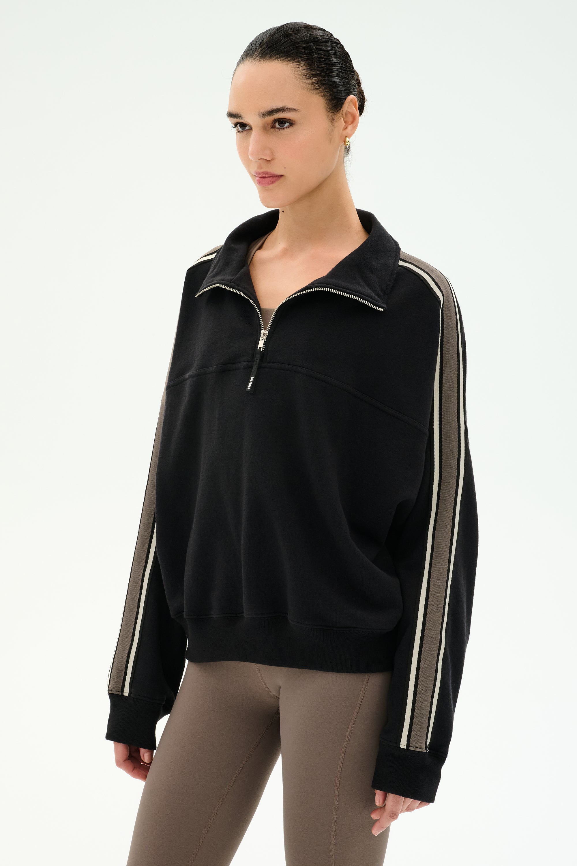 A woman wearing the SPLITS59 Goldie French Terry Oversized Half Zip in Black/Lentil, featuring soft cotton fabric and beige striped sleeves, paired with taupe leggings, stands against a plain white background.