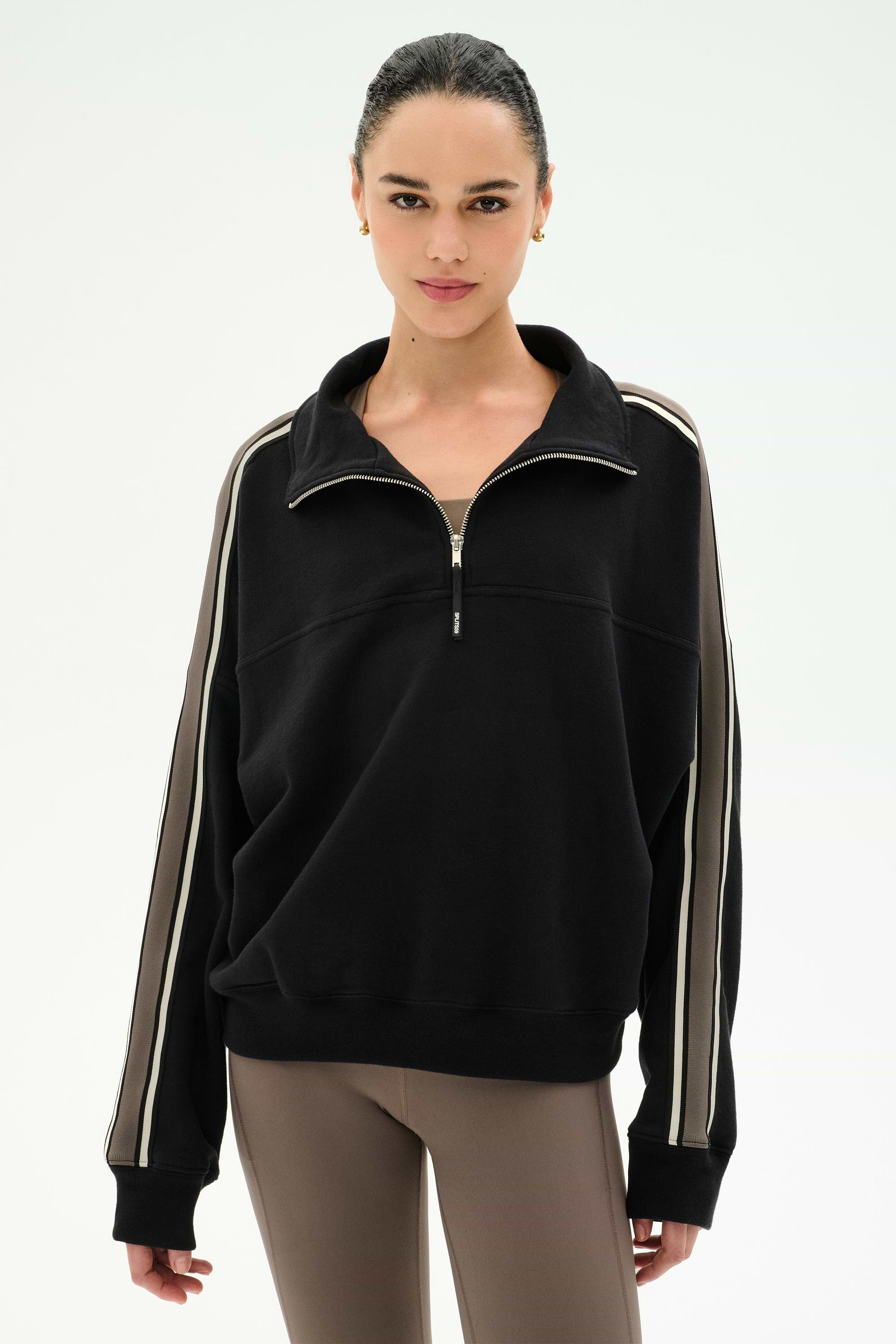 A woman wearing the SPLITS59 Goldie French Terry Oversized Half Zip in Black/Lentil, featuring striped sleeves and soft cotton fabric, paired with taupe leggings, stands against a plain white background.