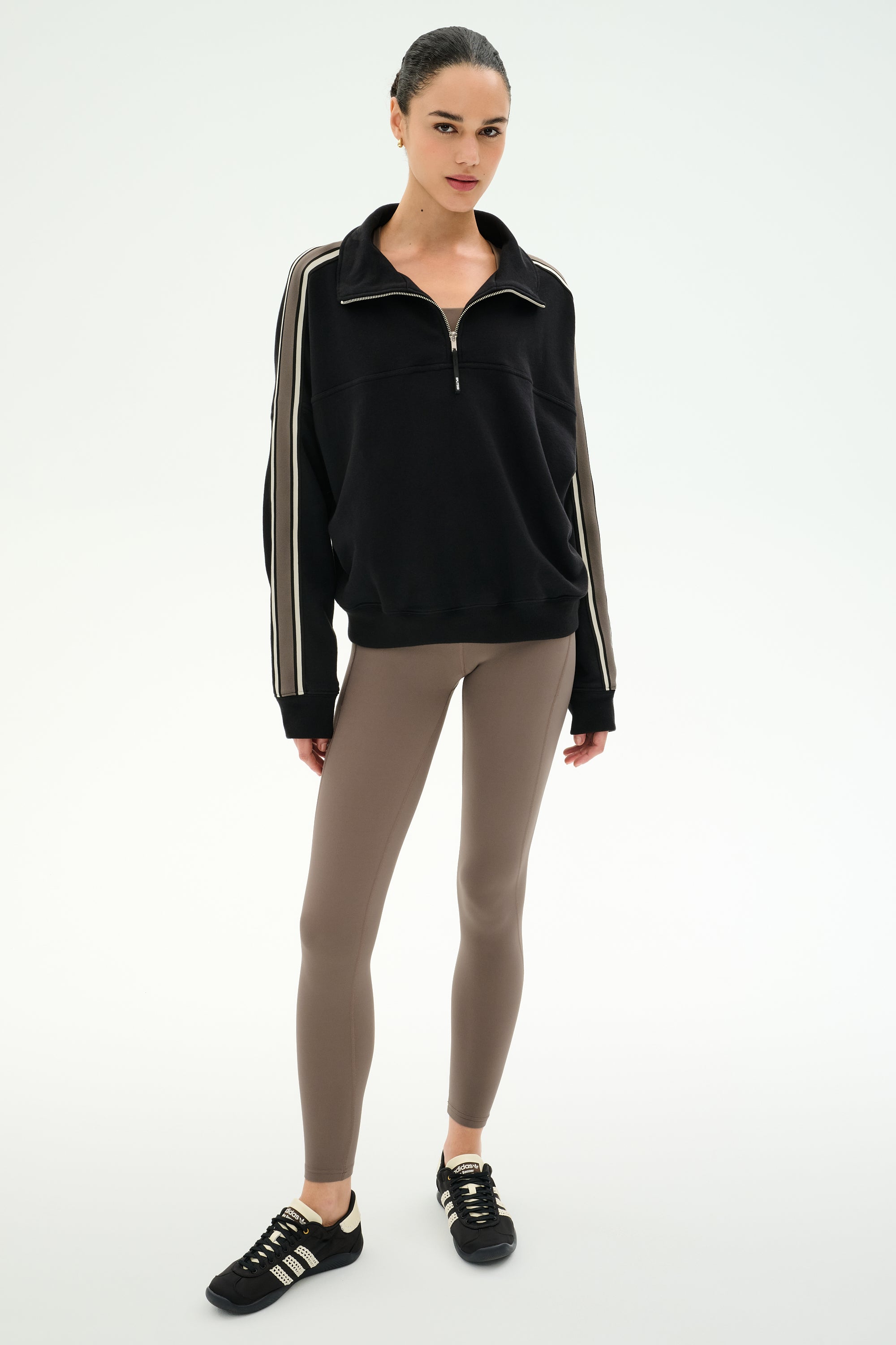 A woman stands against a plain background wearing the SPLITS59 Goldie French Terry Oversized Half Zip in Black/Lentil, featuring soft cotton fabric and striped sleeves, paired with taupe leggings and black sneakers.