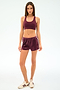 A woman stands facing forward against a plain light background, wearing SPLITS59’s Heritage Velour Short in Dark Cherry/White, a matching maroon sports bra, and white sneakers with maroon stripes.
