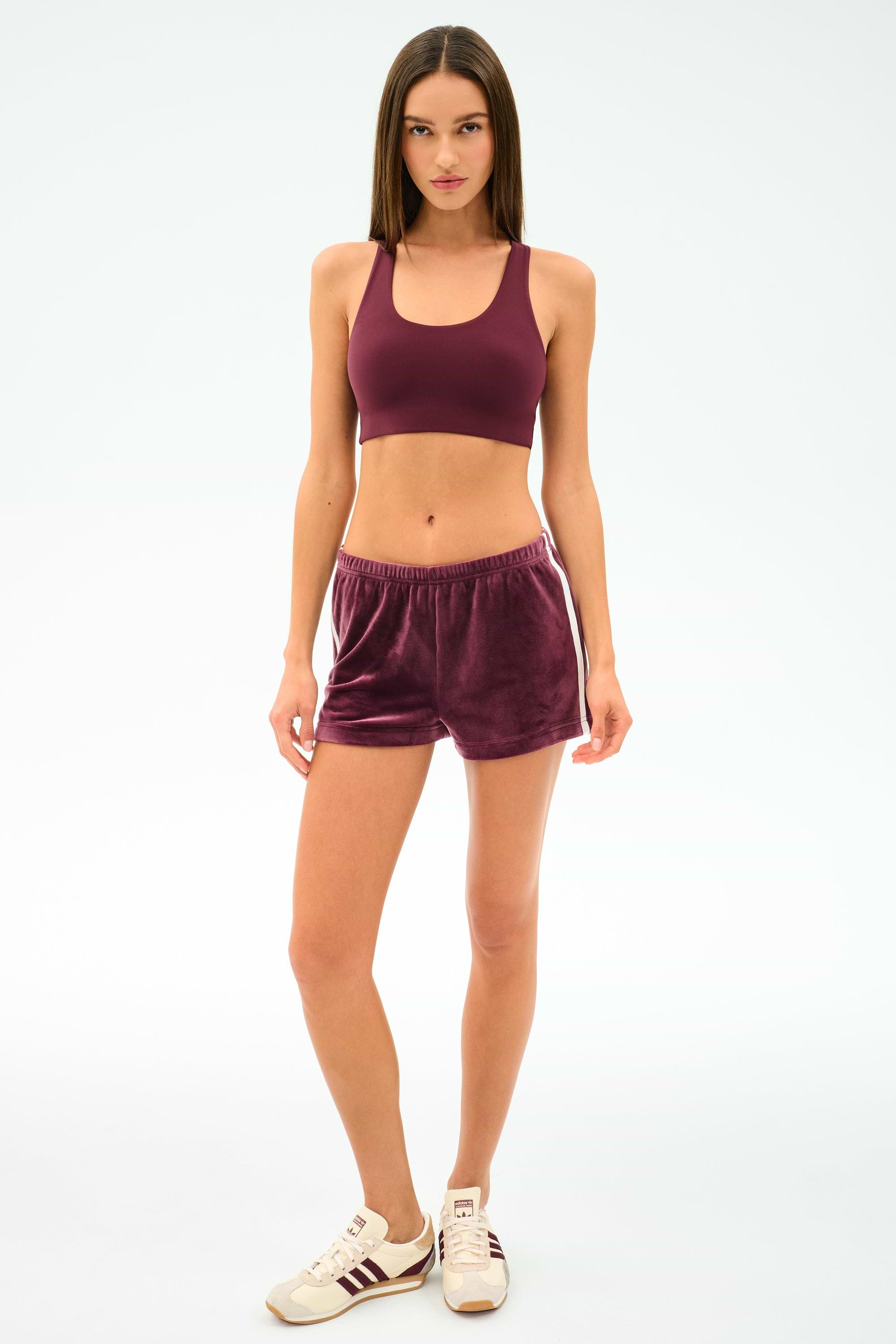 A woman stands facing forward against a plain light background, wearing SPLITS59’s Heritage Velour Short in Dark Cherry/White, a matching maroon sports bra, and white sneakers with maroon stripes.