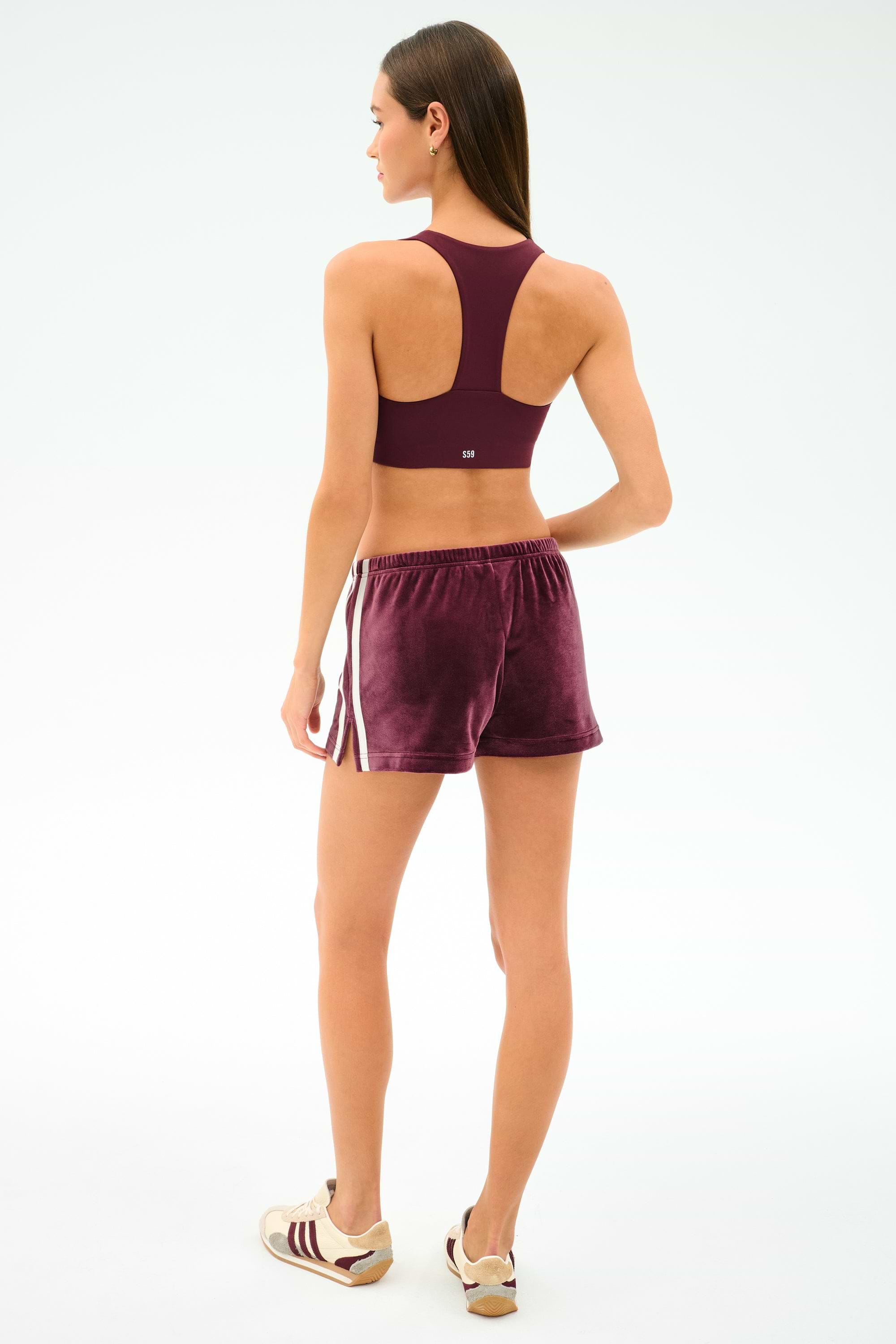 A woman stands facing away, wearing the SPLITS59 Heritage Velour Short in Dark Cherry/White, paired with a burgundy sports bra and sneakers.