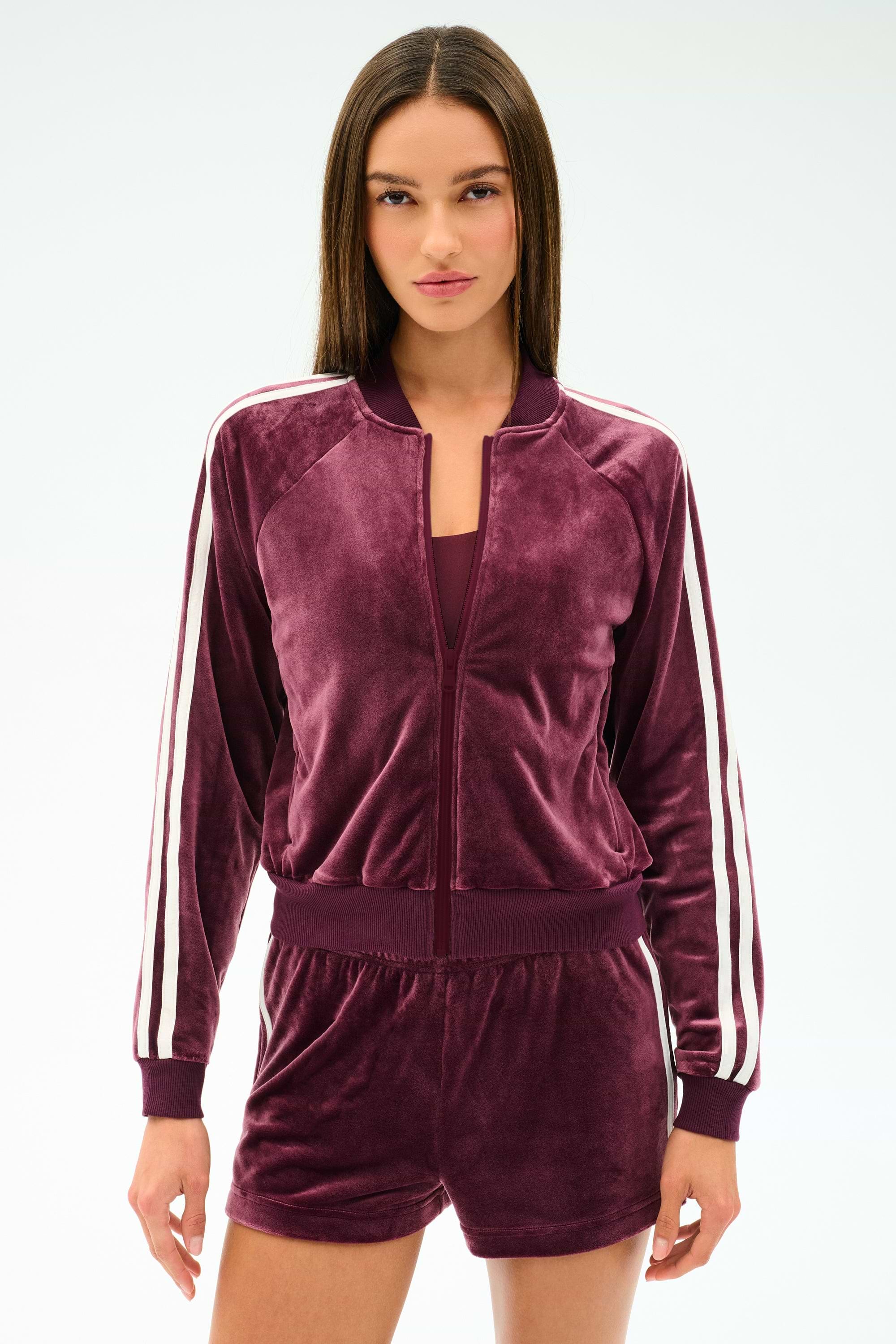 A woman wears the SPLITS59 Heritage Velour Bomber in Dark Cherry/White, featuring white sleeve stripes, paired with matching shorts, standing against a plain light background.