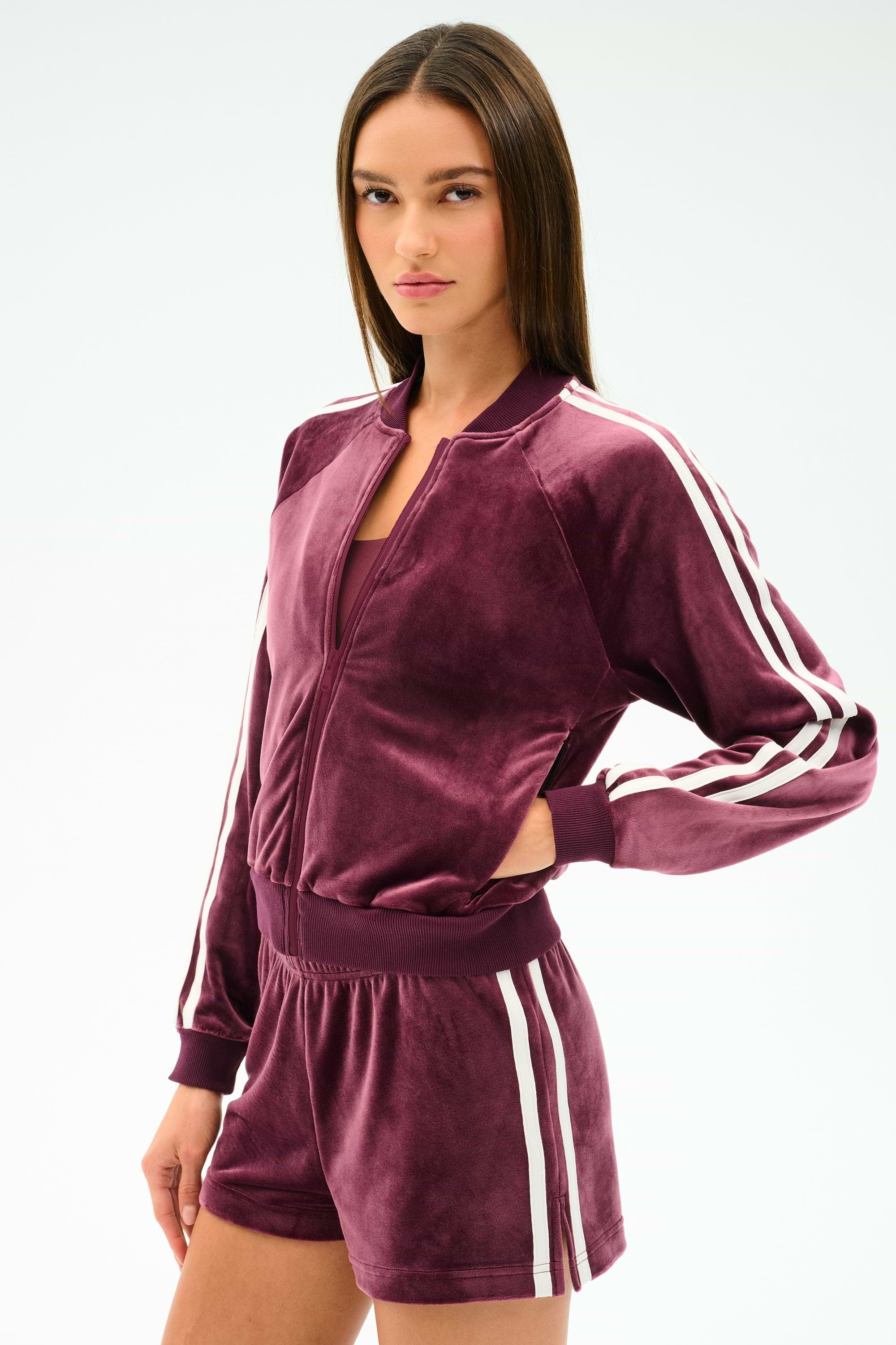 A woman poses with one hand on her waist, wearing the SPLITS59 Heritage Velour Bomber in Dark Cherry/White, paired with matching shorts featuring white side stripes, set against a plain background.