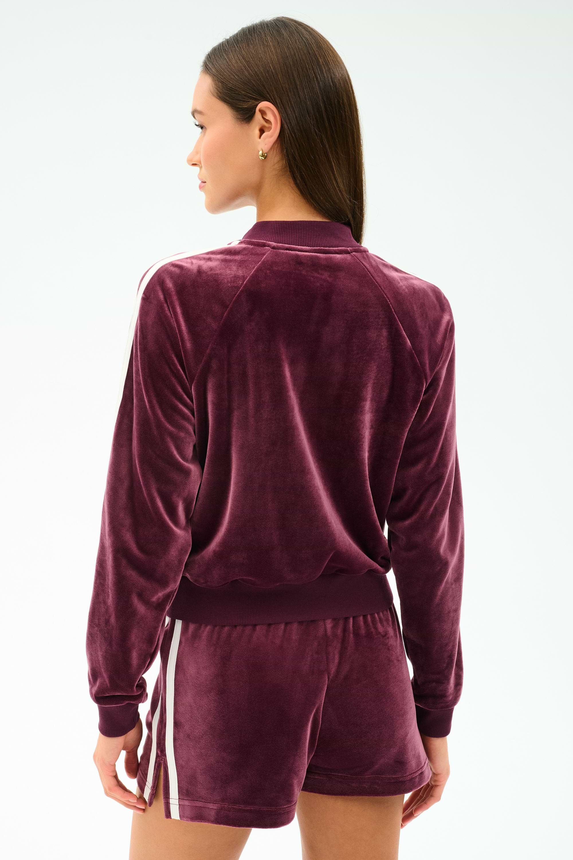 A woman stands with her back to the camera, wearing the SPLITS59 Heritage Velour Bomber in Dark Cherry/White, featuring white sleeve stripes and shorts, made from a soft polyester-spandex blend for comfort and flexibility.