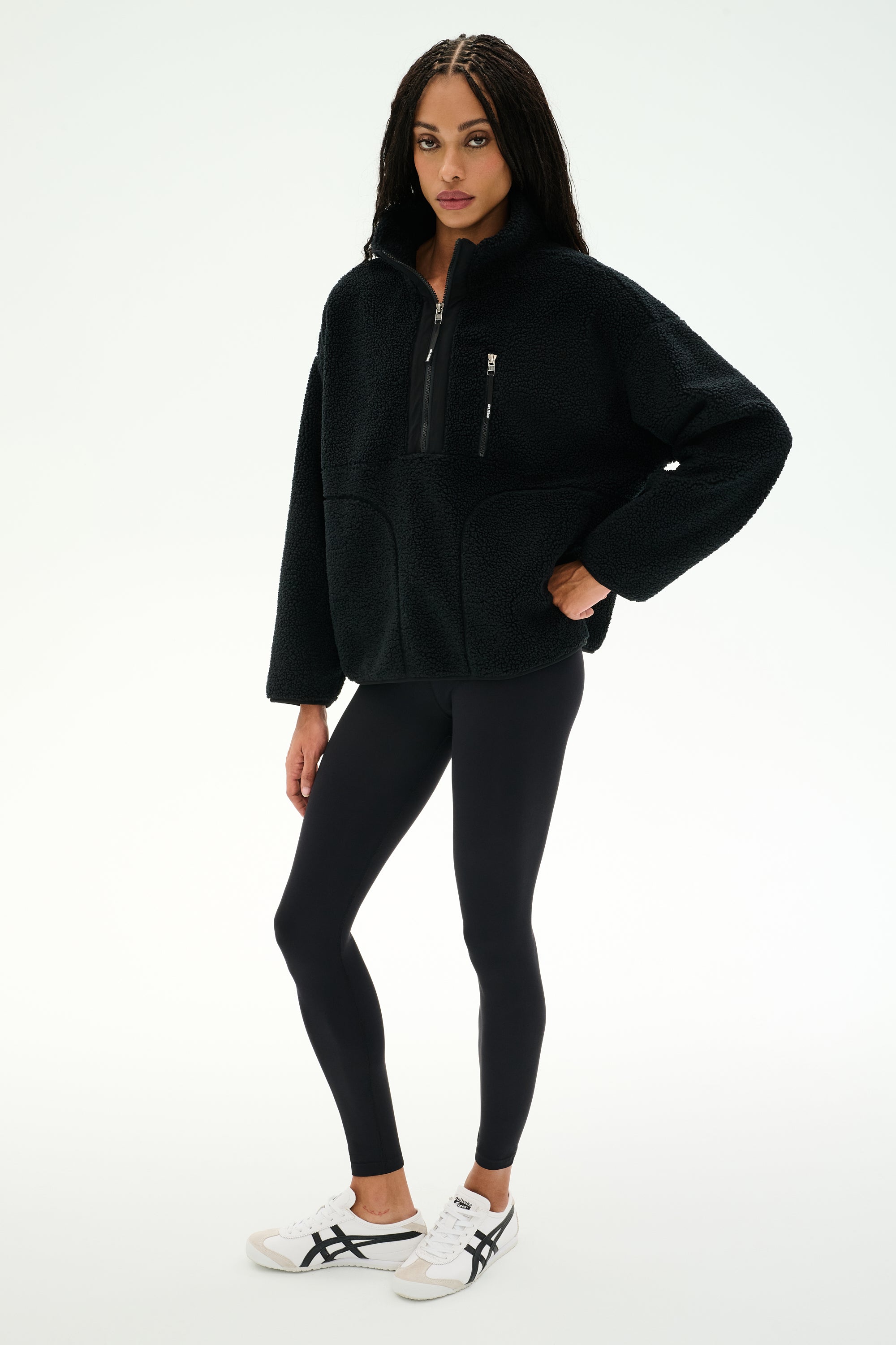 A woman wears the SPLITS59 Reese Sherpa Half Zip Pullover in black, styled with black leggings and white sneakers, standing against a plain white background.