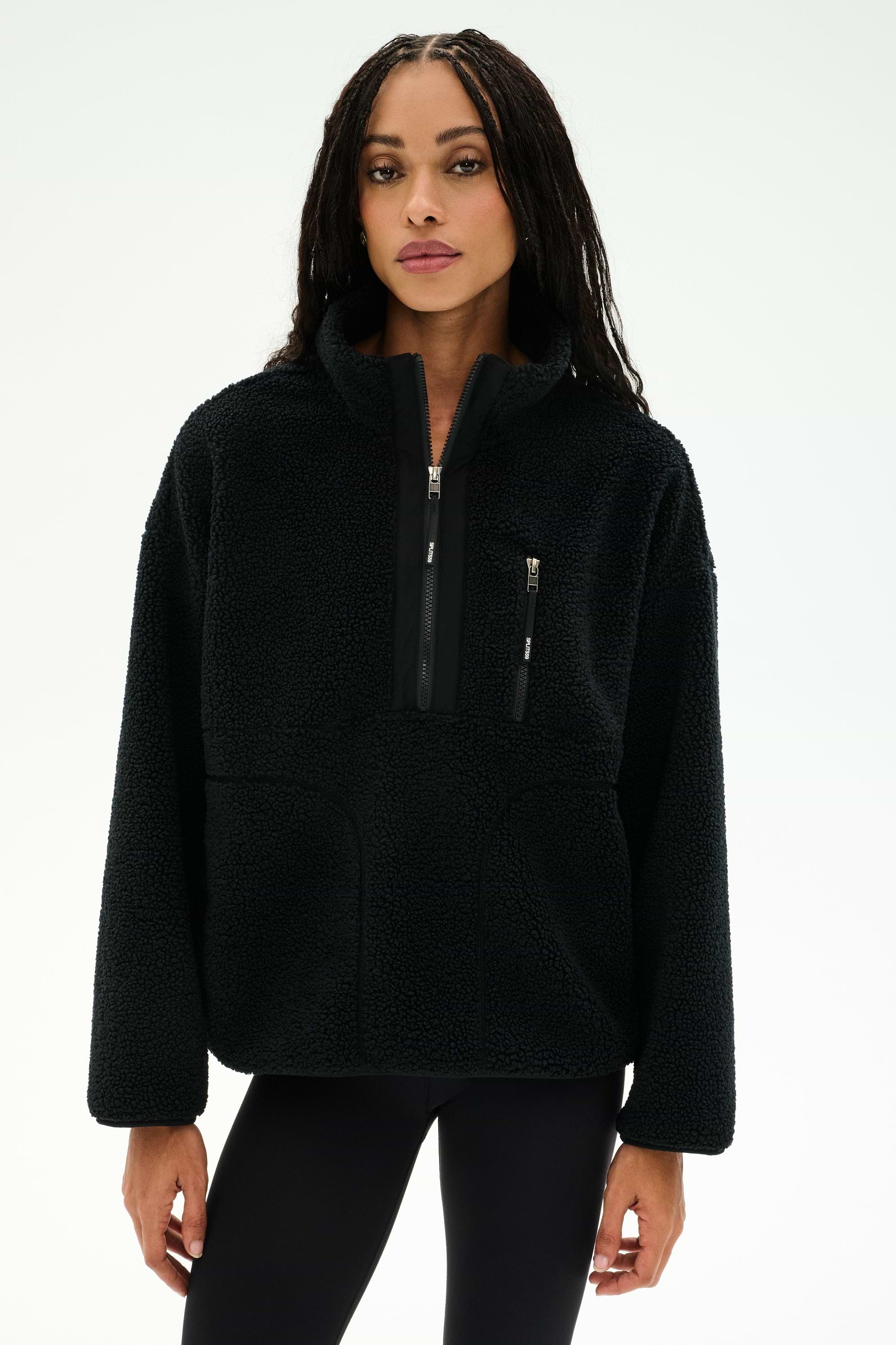 A woman wearing the SPLITS59 Reese Sherpa Half Zip Pullover in black, a soft polyester fleece with long sleeves and front pockets, stands against a plain white background.