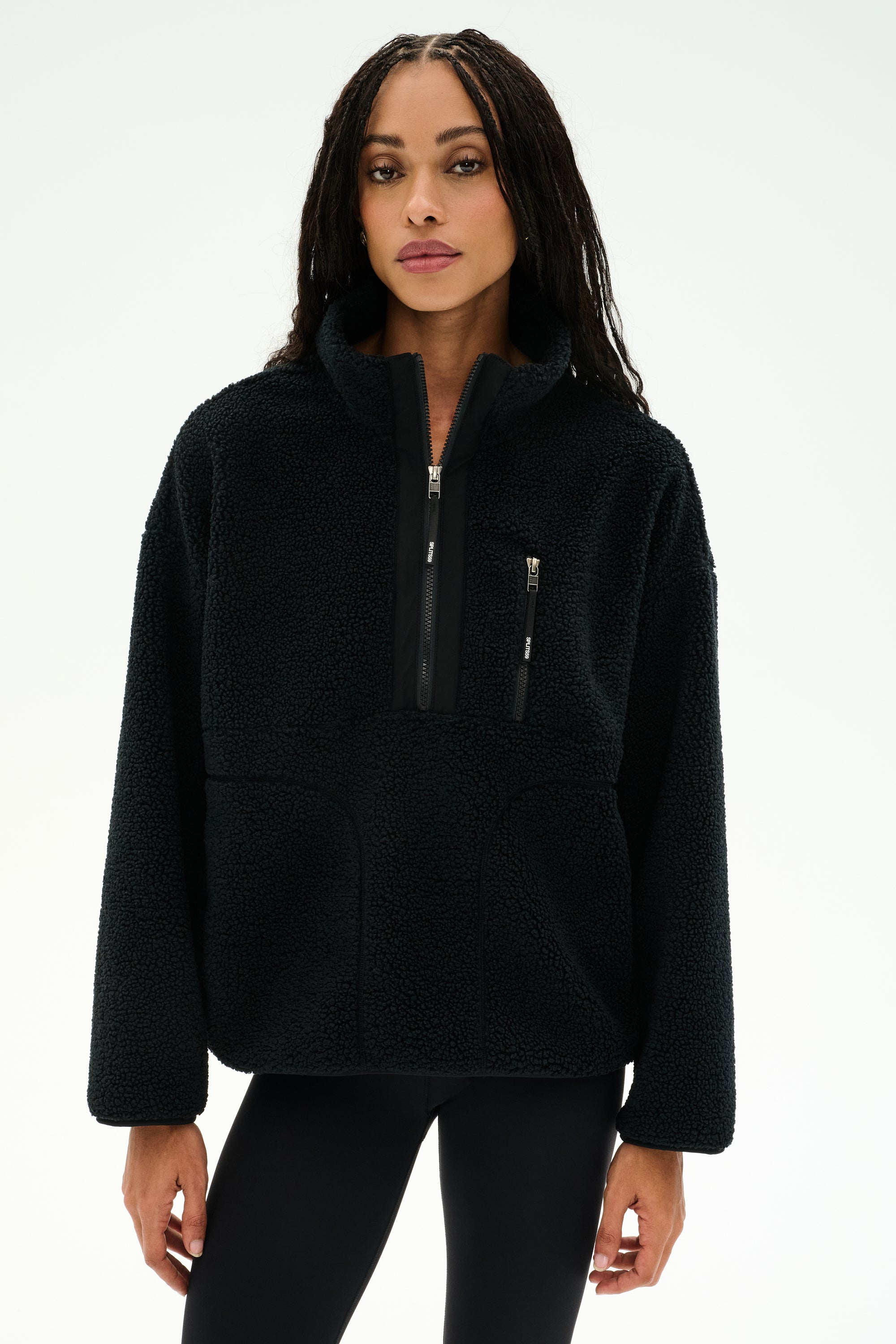 A woman wearing the SPLITS59 Reese Sherpa Half Zip Pullover in black, a soft polyester fleece with long sleeves and front pockets, stands against a plain white background.
