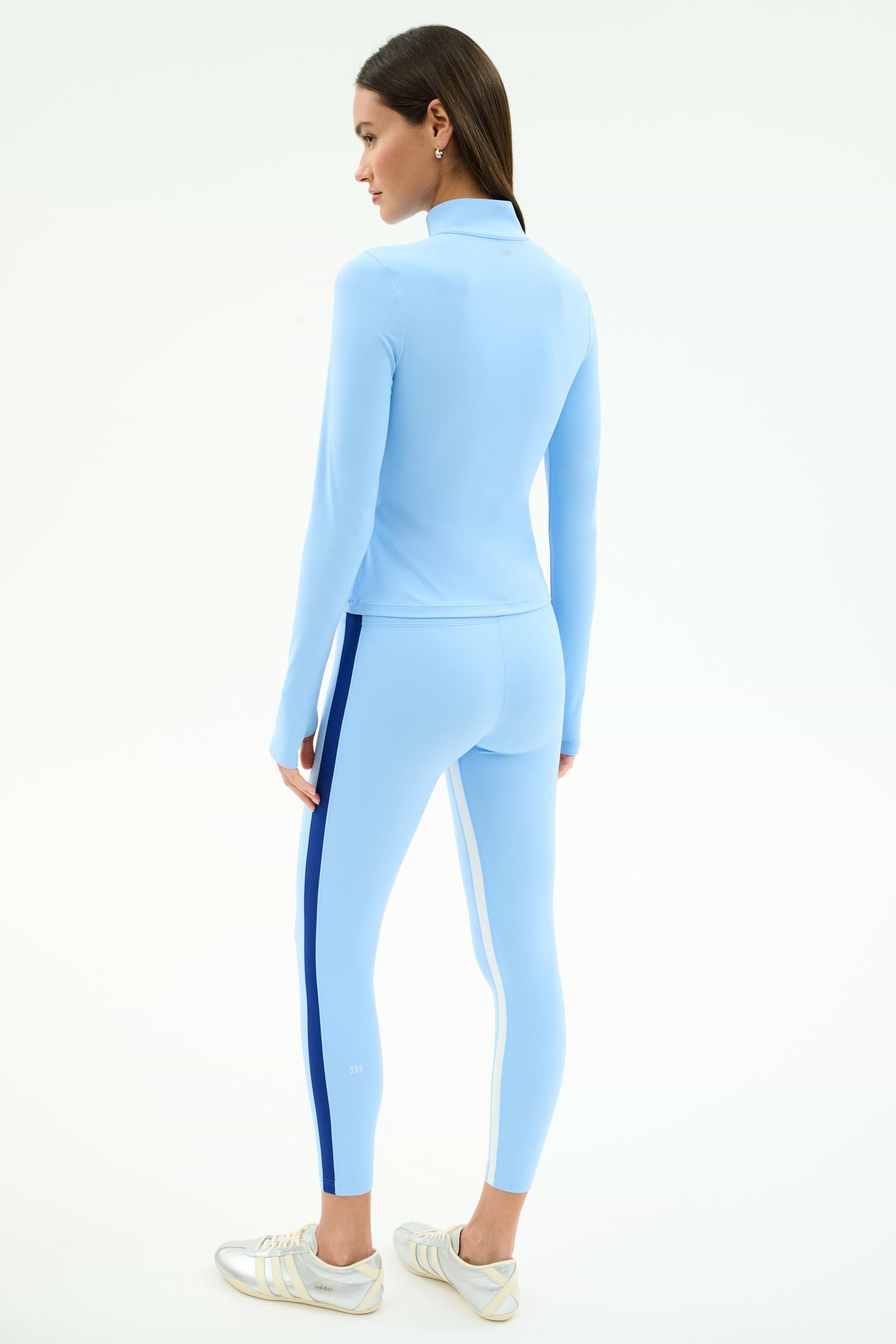 A woman wearing the SPLITS59 Tess Rigor Half Zip Waist Length jacket in Skylight with matching leggings and white sneakers, standing and facing away on a plain background.