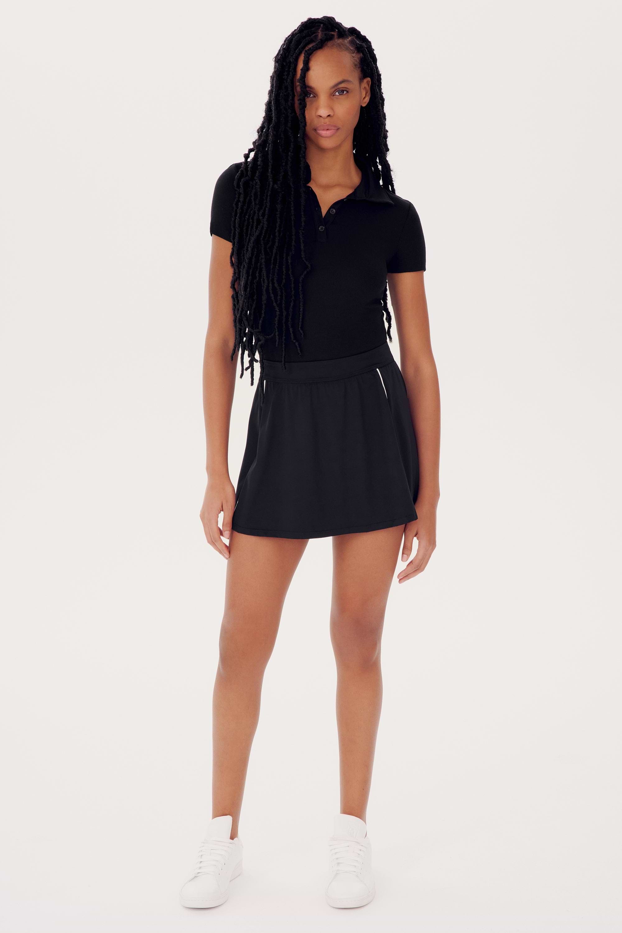 A young woman in a Talia Rib Polo - Black by SPLITS59 and skirt poses while wearing white sneakers, standing against a plain white background.