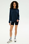 A woman faces forward in a SPLITS59 Andie Fleece Sweatshirt - Indigo, black shorts with a red stripe, white athletic socks, and gray sneakers against a plain light background.
