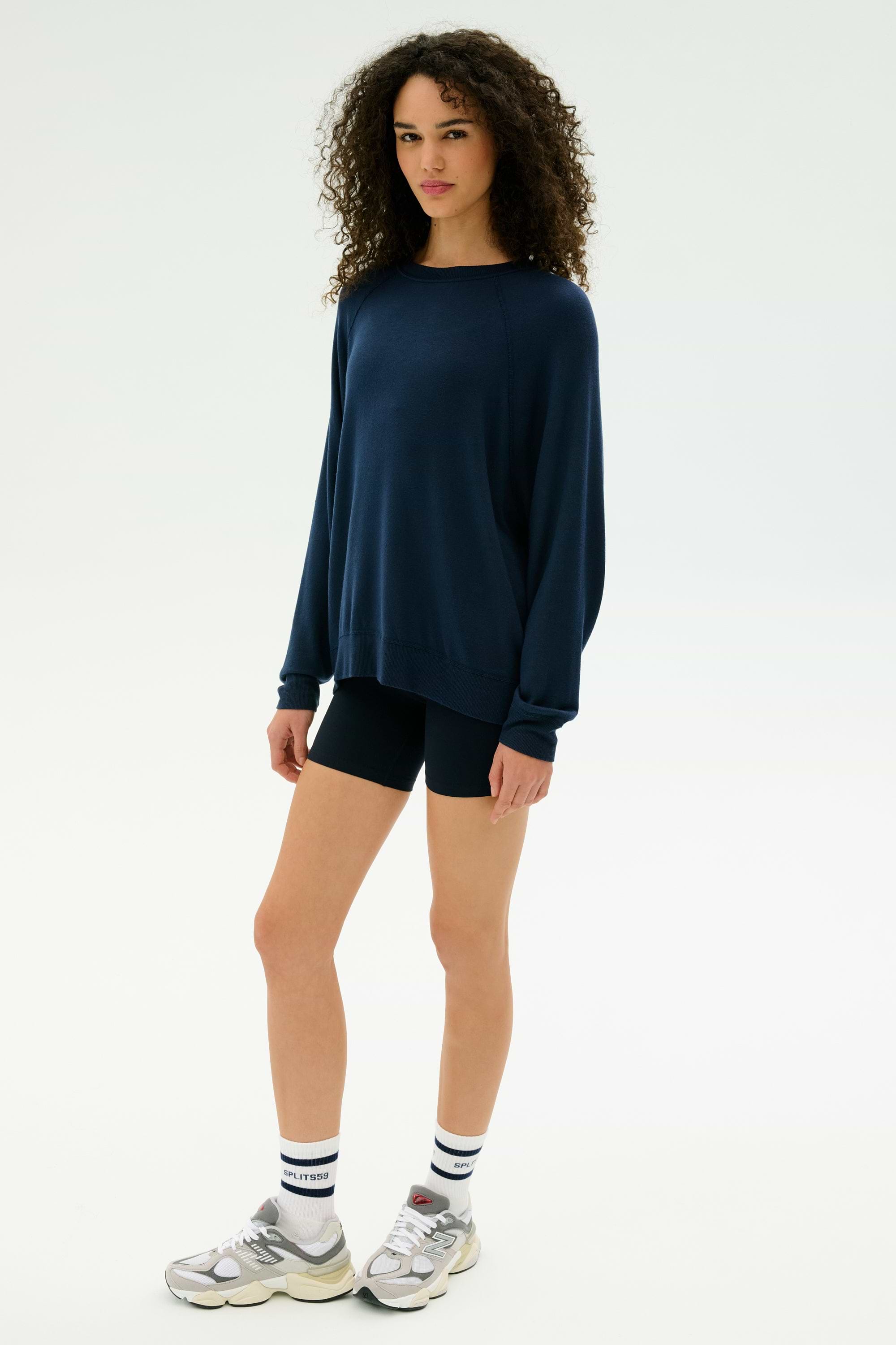 A person stands against a plain background wearing the SPLITS59 Andie Fleece Sweatshirt in Indigo, paired with black shorts, white socks, and silver sneakers.