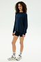A person stands against a plain background wearing the SPLITS59 Andie Fleece Sweatshirt in Indigo, paired with black shorts, white socks, and silver sneakers.