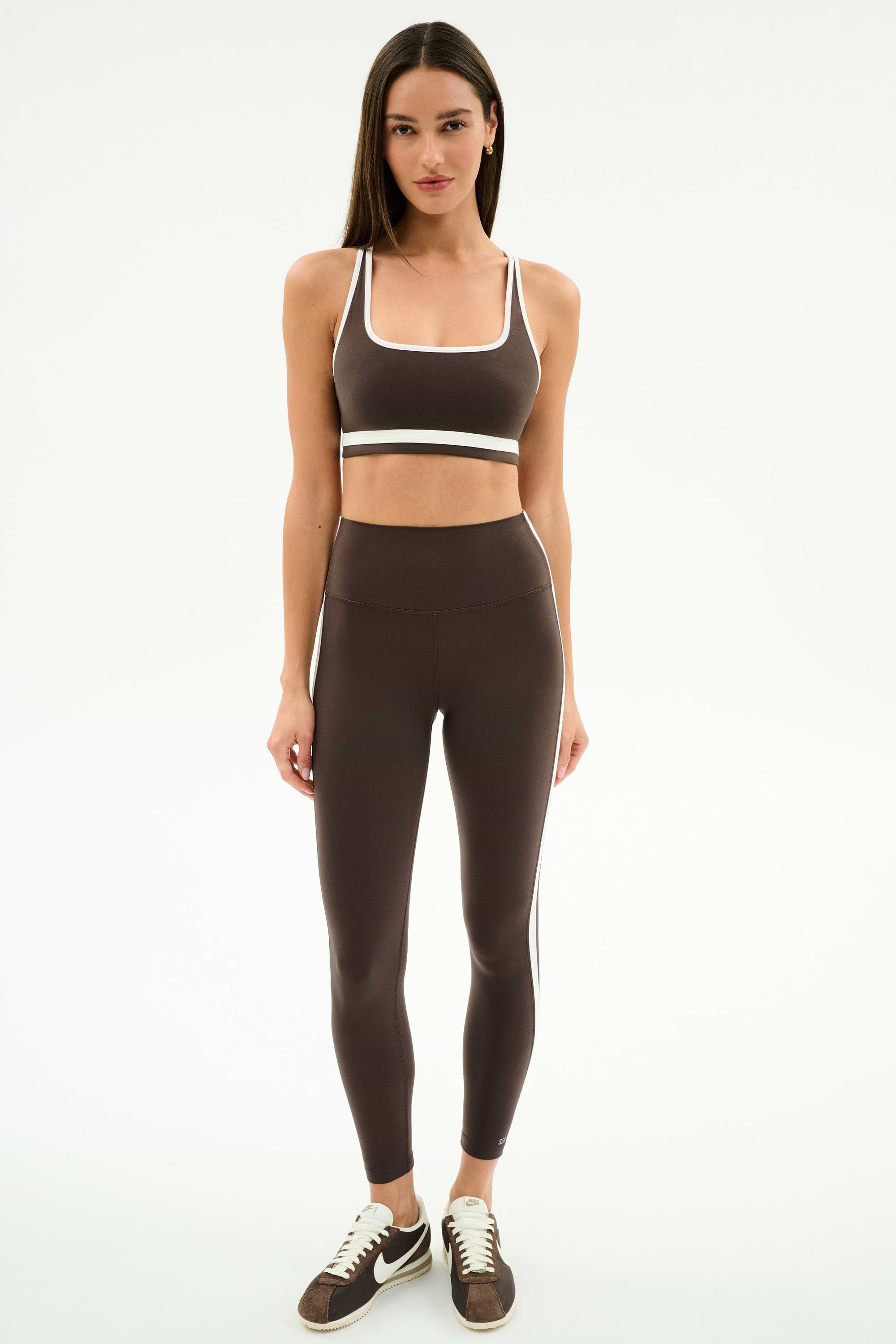A woman stands facing forward against a white background, wearing SPLITS59's Bianca High Waist Rigor 7/8 leggings in Dark Chocolate/White and a matching sports bra.