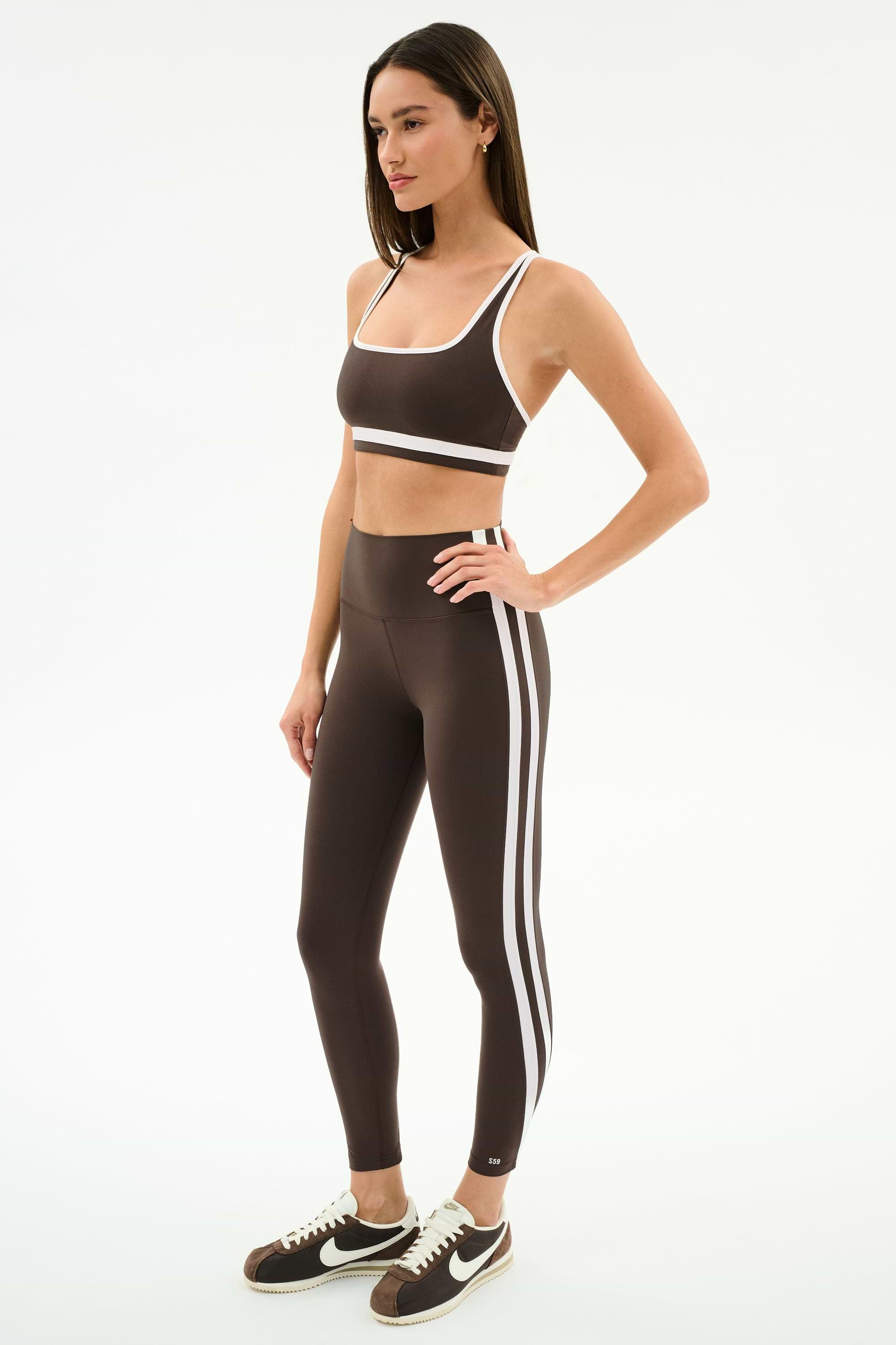 A woman wears the SPLITS59 Bianca High Waist Rigor 7/8 leggings in Dark Chocolate/White, featuring white side stripes and performance fabric, paired with a matching sports bra and black-and-white sneakers.