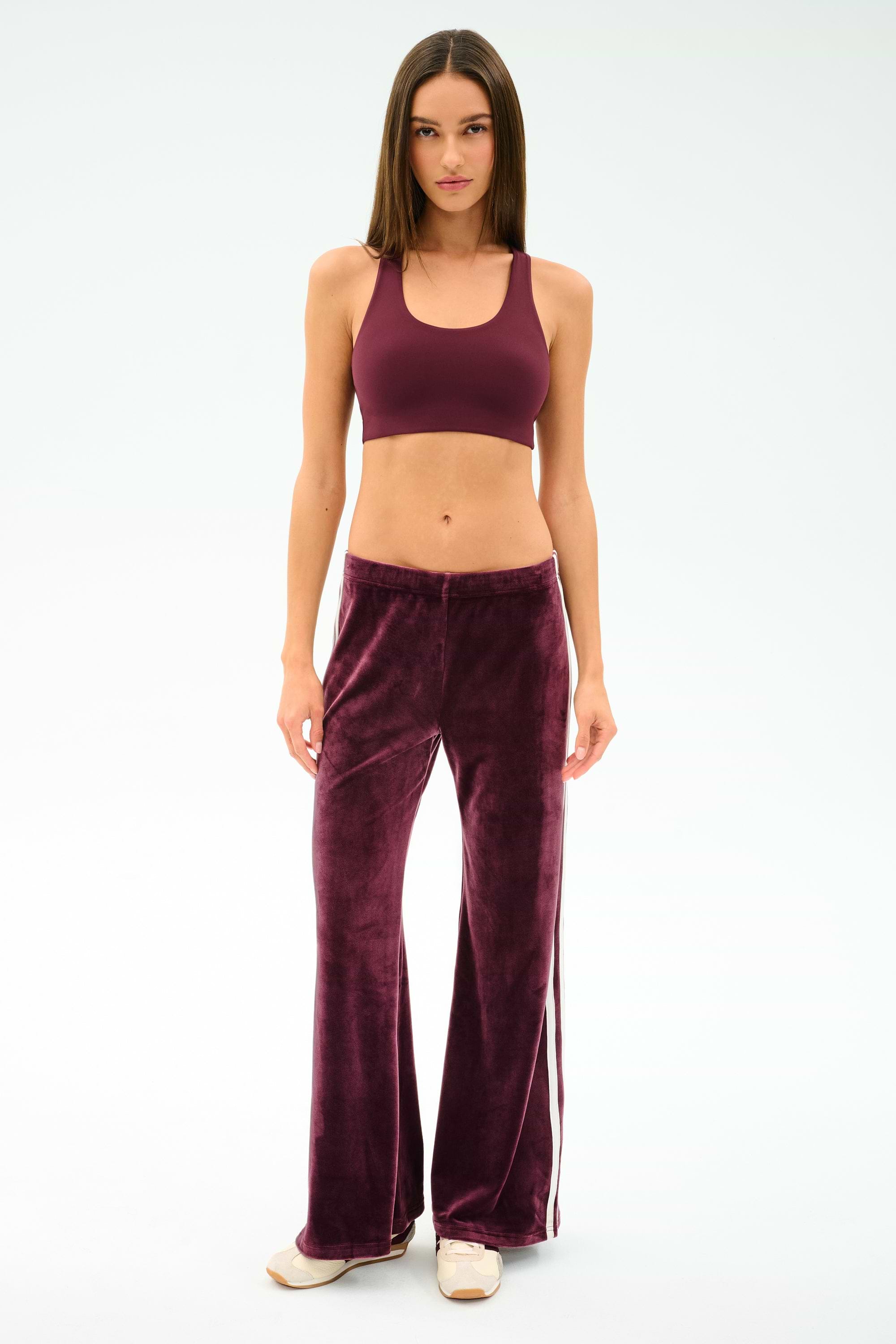 A woman stands facing forward in SPLITS59 Heritage Velour Pant in Dark Cherry/White, paired with a matching burgundy sports bra and beige shoes, set against a plain white background.
