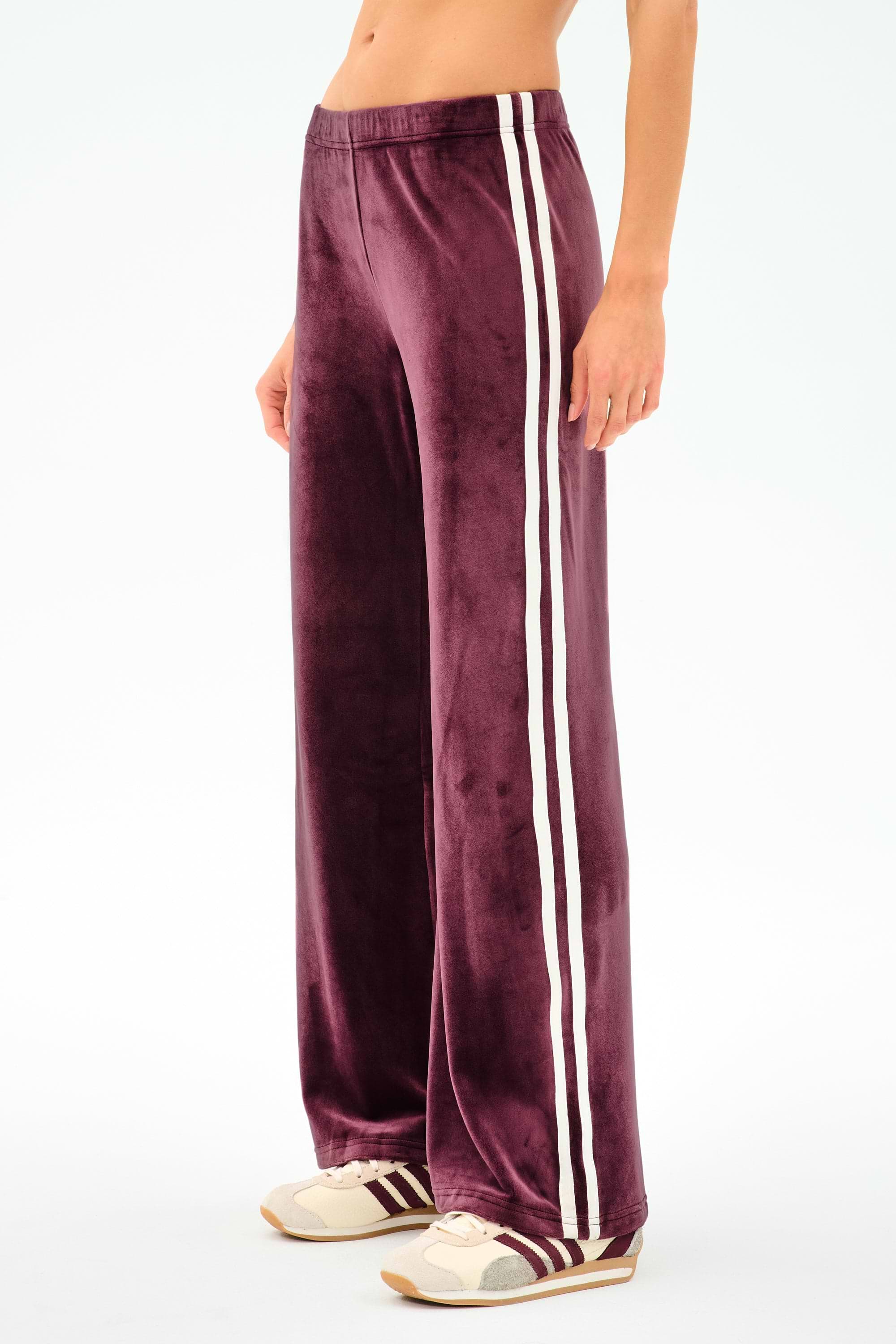 A person wears SPLITS59 Heritage Velour Pant in Dark Cherry/White, featuring wide legs, white side stripes, and a stretchy spandex blend, paired with beige sneakers—shown waist down against a white background.