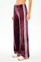 A person wears SPLITS59 Heritage Velour Pant in Dark Cherry/White, featuring wide legs, white side stripes, and a stretchy spandex blend, paired with beige sneakers—shown waist down against a white background.