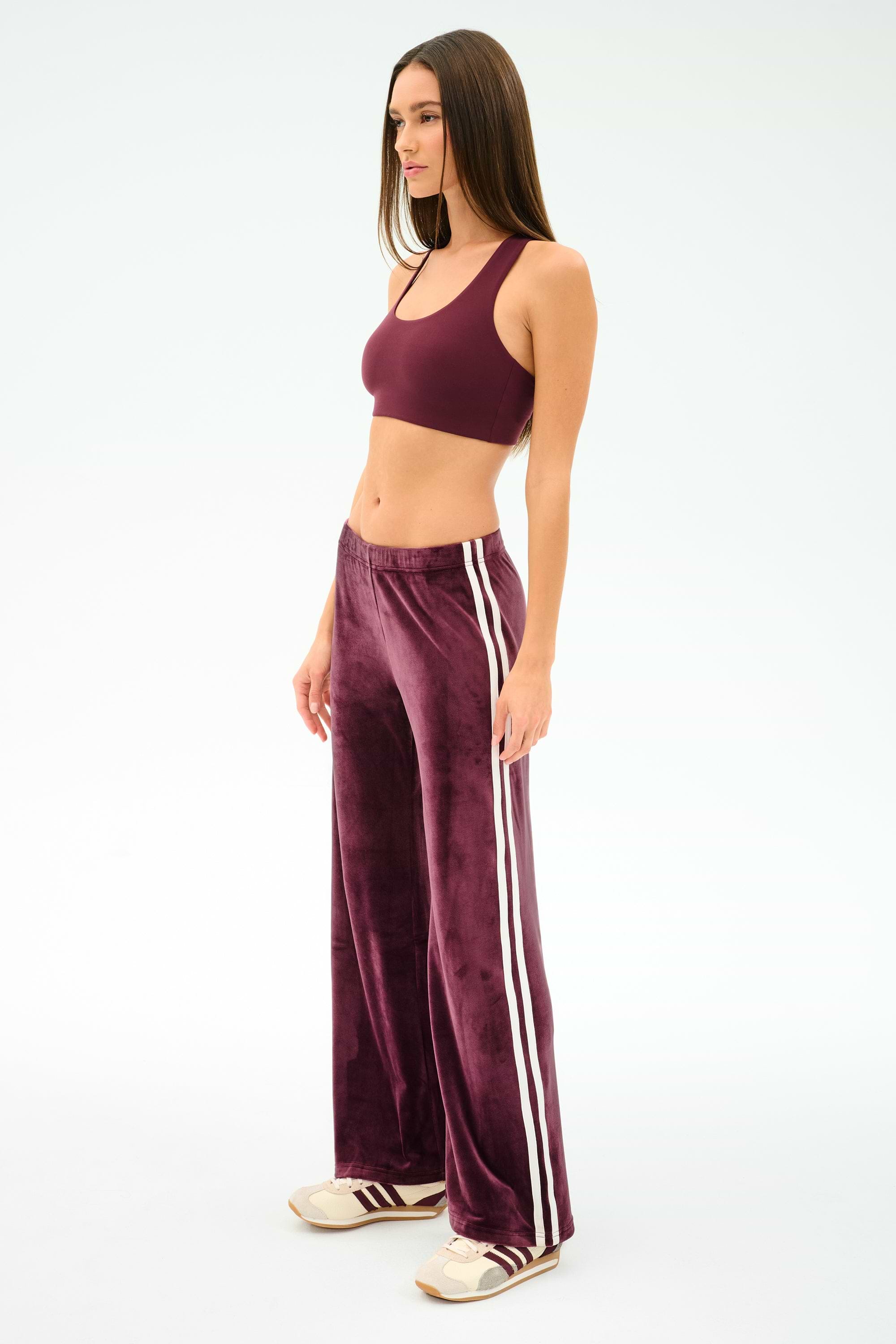 A woman in profile wears SPLITS59’s Heritage Velour Pant in Dark Cherry/White, made from stretchy velour with white stripes, paired with a burgundy sports bra and white sneakers against a plain background.