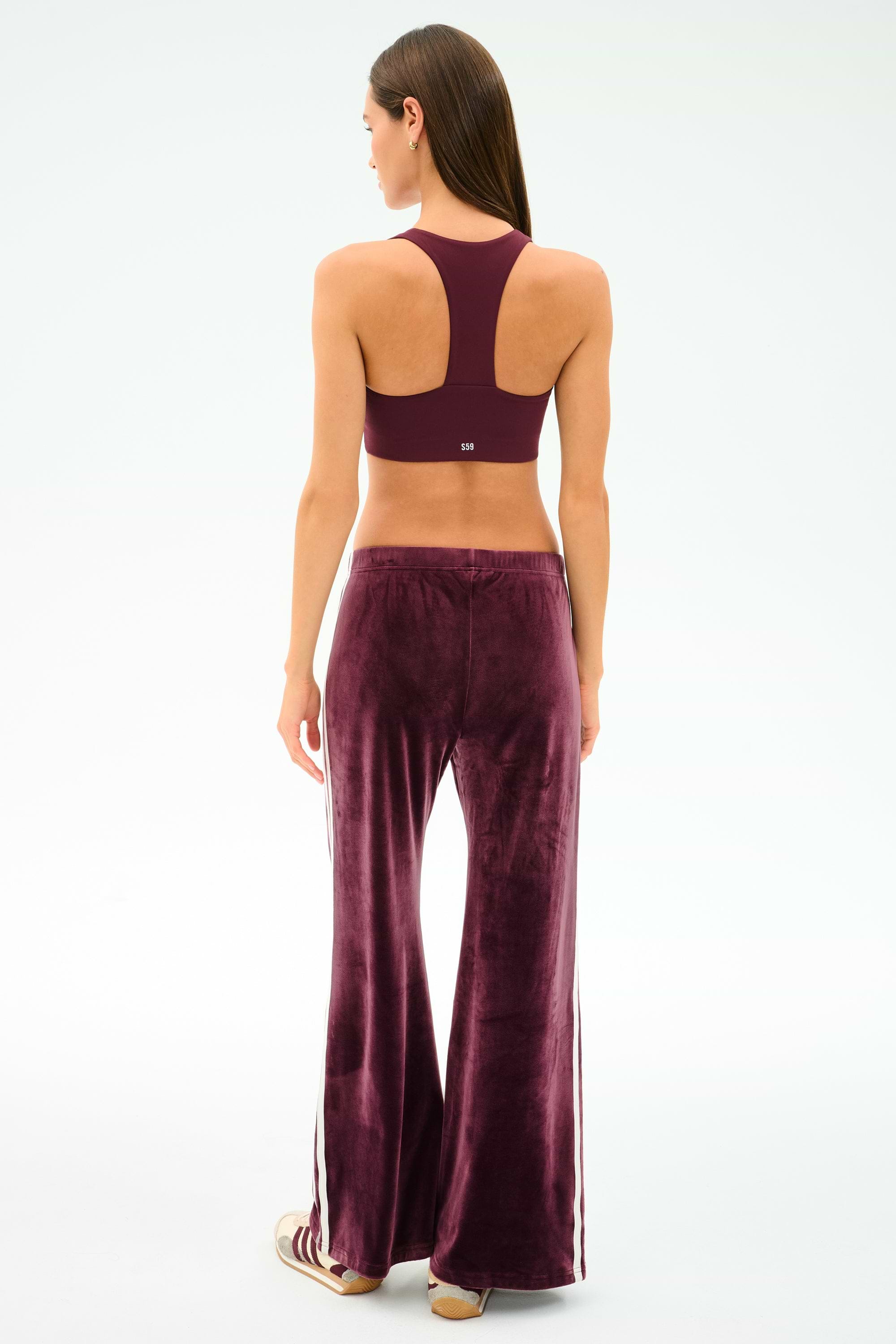 A woman stands facing away, wearing SPLITS59 Heritage Velour Pant in Dark Cherry/White with side stripes and a matching maroon racerback sports bra, crafted from a stretchy spandex blend, against a plain light background.