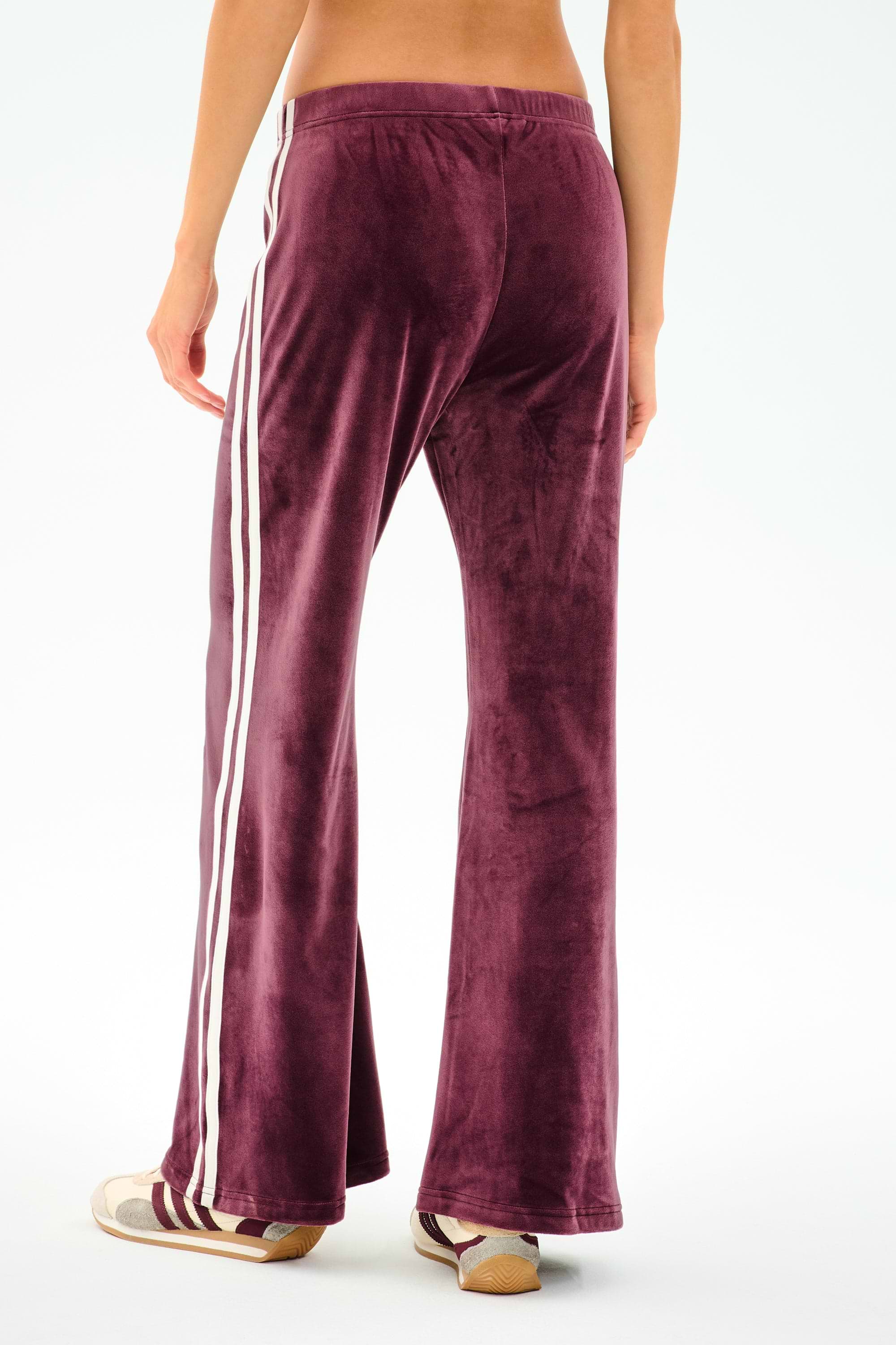 A model wears SPLITS59 Heritage Velour Pant in Dark Cherry/White, made from a soft polyester-spandex blend, with white side stripes and beige sneakers, shown from the back against a plain white background.