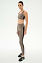 A woman stands in a neutral pose against a plain white background, wearing the SPLITS59 Margot Rigor Legging in Lentil/Black—a taupe legging with black stripe detail crafted from a soft 62% nylon, 38% spandex blend—paired with a matching sports bra and black sneakers.