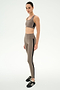 A woman stands in a neutral pose against a plain white background, wearing the SPLITS59 Margot Rigor Legging in Lentil/Black—a taupe legging with black stripe detail crafted from a soft 62% nylon, 38% spandex blend—paired with a matching sports bra and black sneakers.