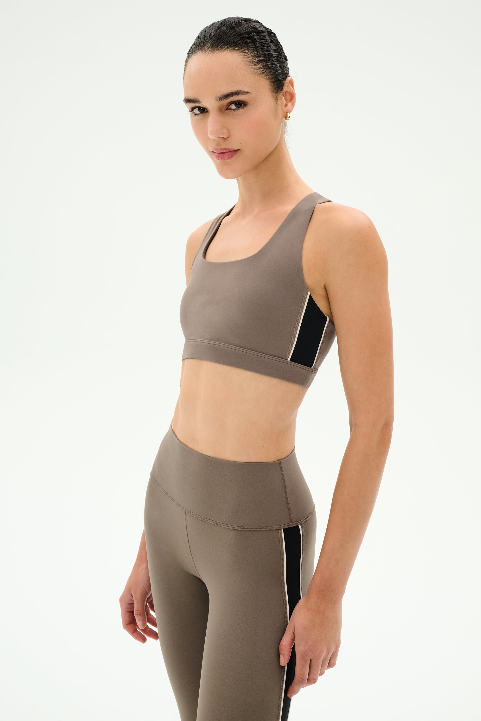 A woman wearing the SPLITS59 Lexi Rigor Bra in Lentil/Black, paired with matching high-waisted leggings featuring black side accents. Both pieces are crafted from soft stretch fabric for ultimate comfort, as she stands against a plain white background.