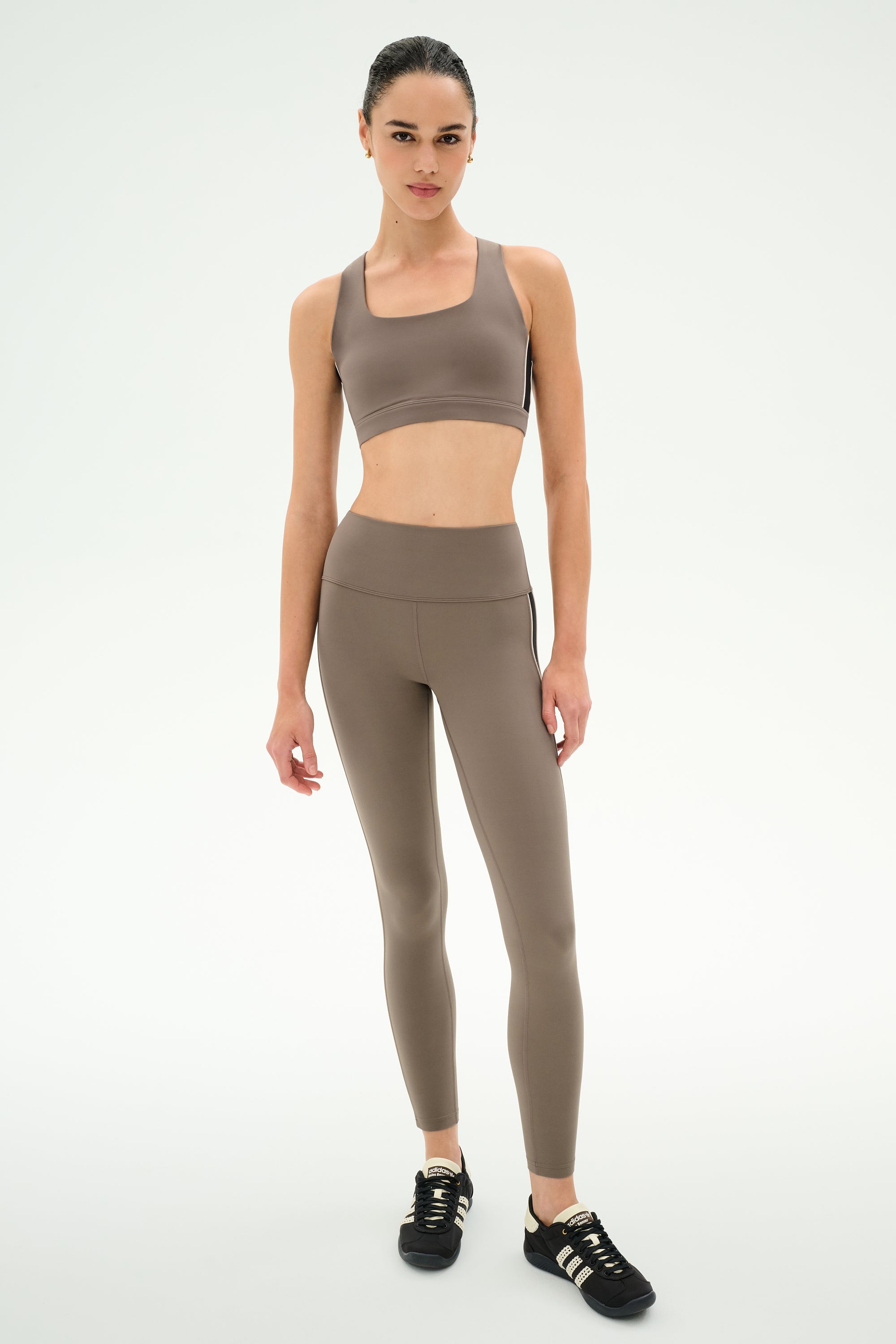 Model stands against a plain background wearing the SPLITS59 Lexi Rigor Bra in Lentil/Black, paired with matching high-waisted leggings crafted from stretch fabric and black sneakers—an effortlessly stylish activewear look.
