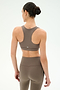 A woman with dark hair in a bun stands with her back to the camera against a plain light background, wearing the SPLITS59 Lexi Rigor Bra in Lentil/Black and matching stretch leggings.