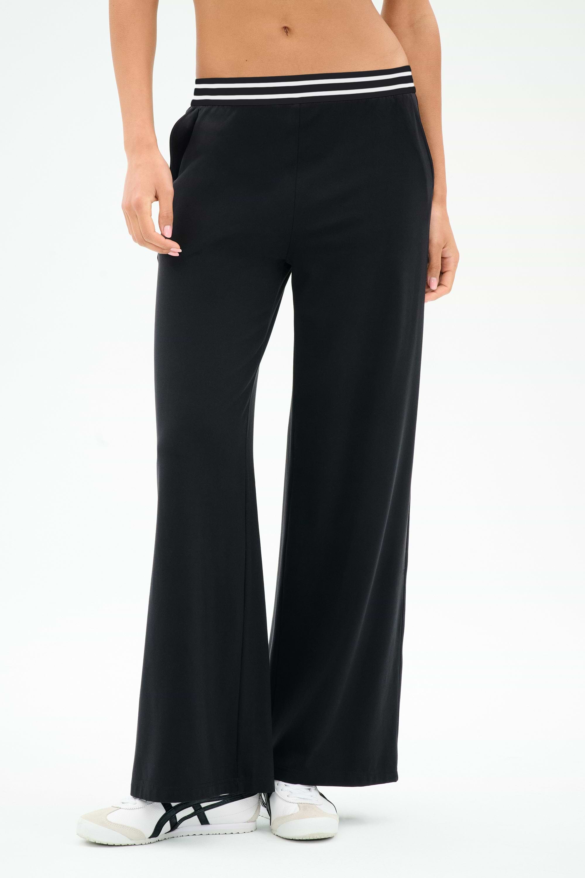 A person wears SPLITS59 Michele Airweight Trouser - Black, featuring a wide-leg fit and a white-and-black striped waistband, paired with white sneakers against a plain background.
