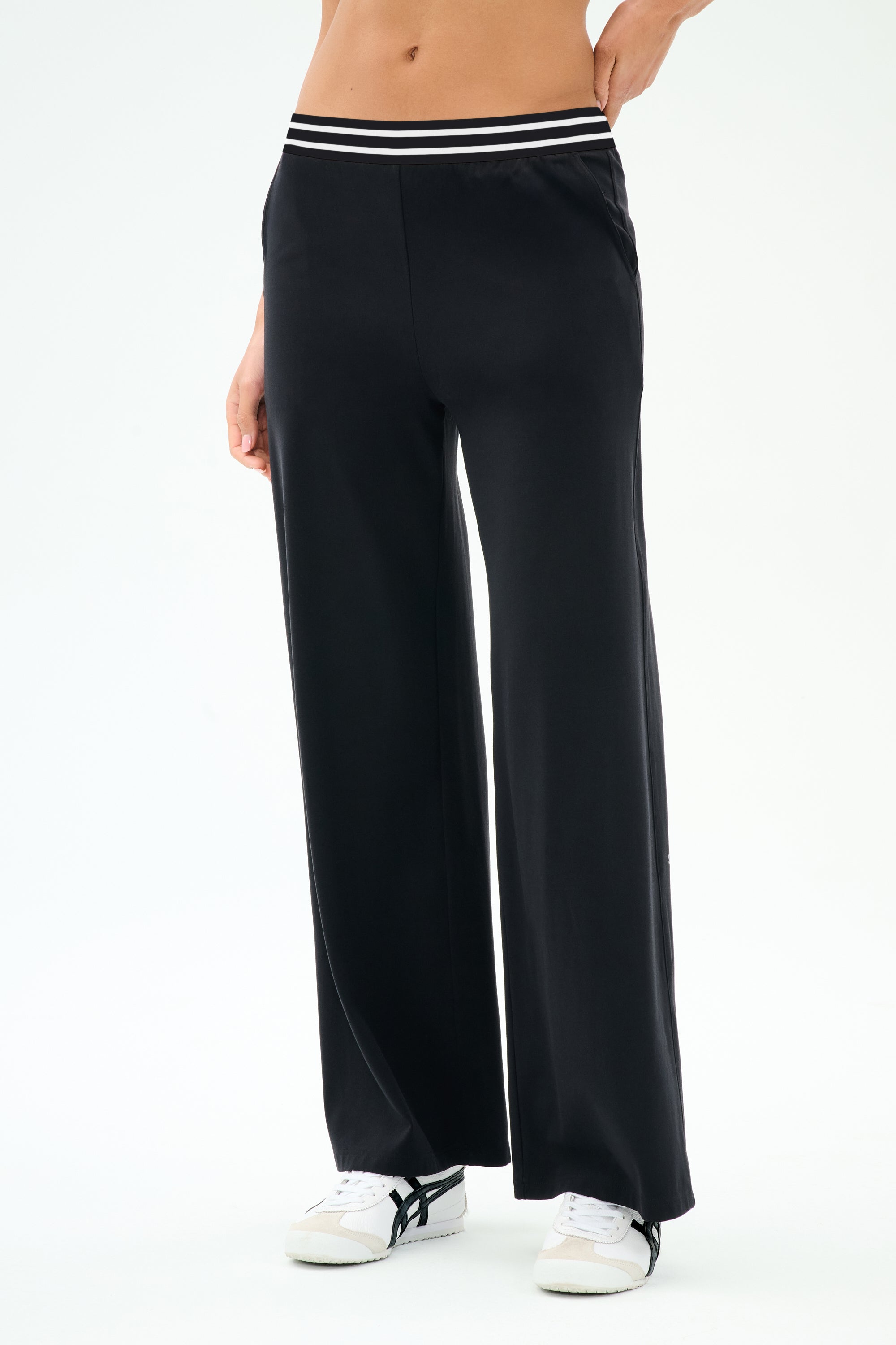 A person wears the SPLITS59 Michele Airweight Trouser in black, featuring a wide-leg cut and a white and black striped waistband, paired with white sneakers, standing against a plain light background.