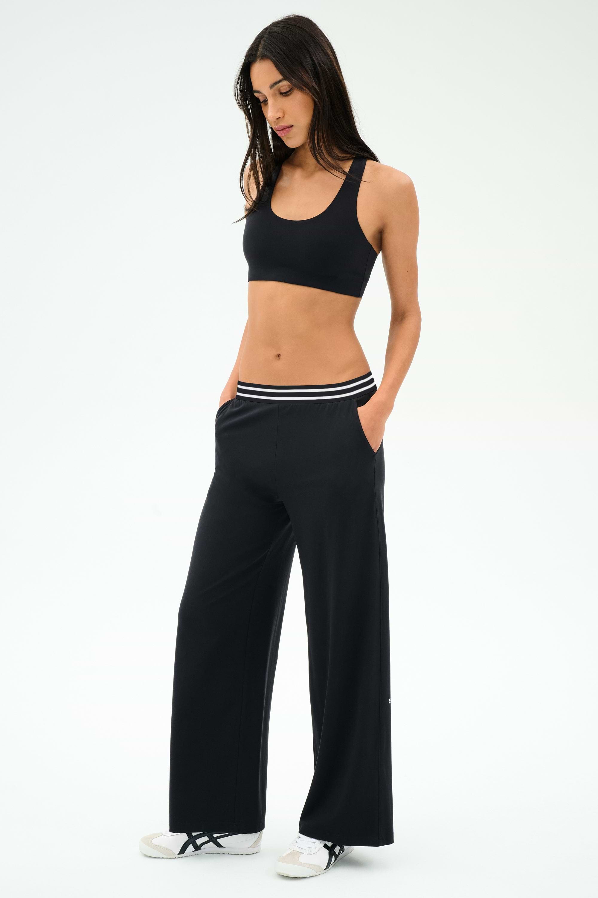 A woman stands with hands in pockets against a plain white background, wearing a black sports bra, SPLITS59 Michele Airweight Trouser - Black with a striped waistband, and white sneakers.