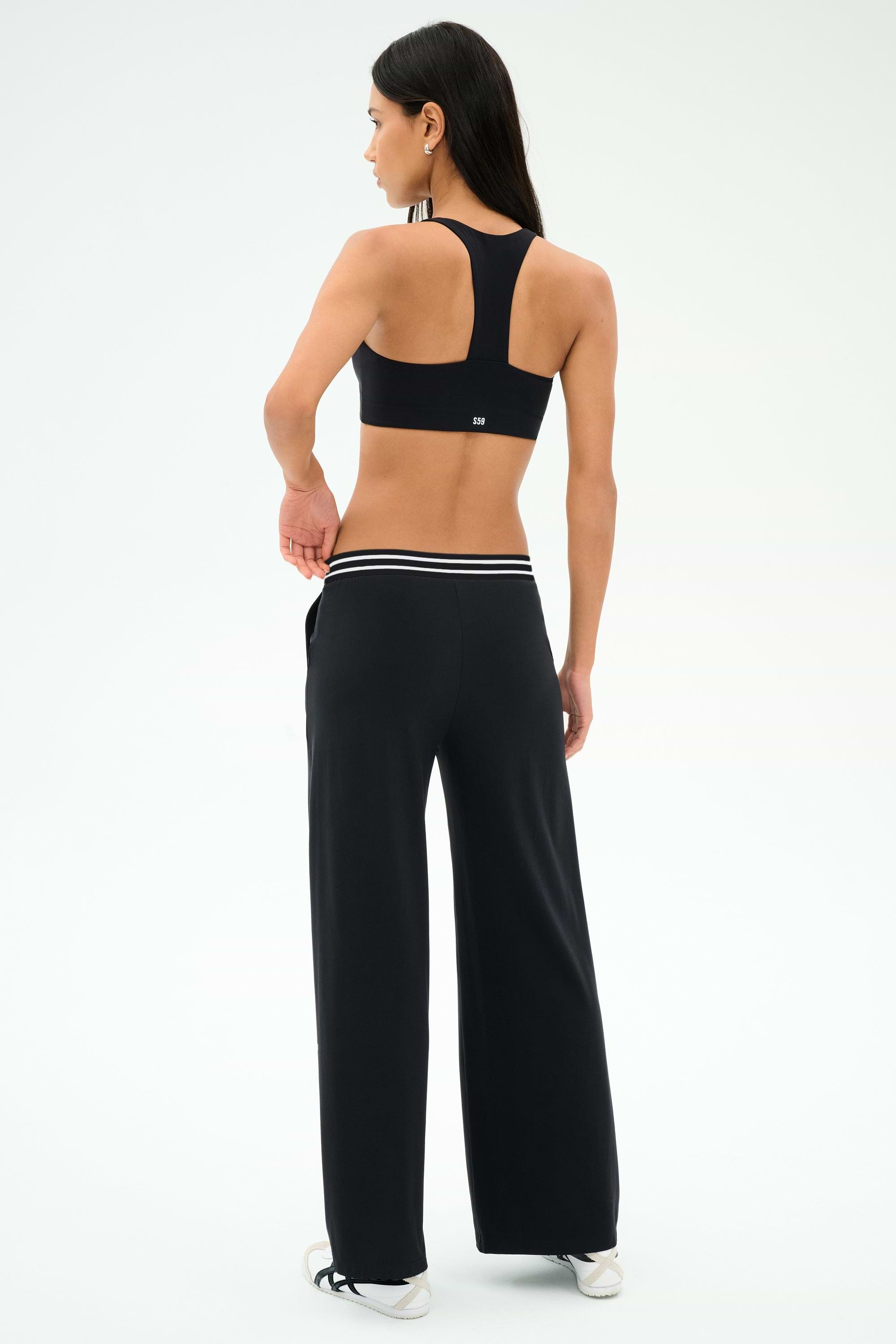 A woman stands facing away, wearing a black sports bra, white sneakers, and SPLITS59 Michele Airweight Trouser - Black with a striped waistband, posed against a white background.