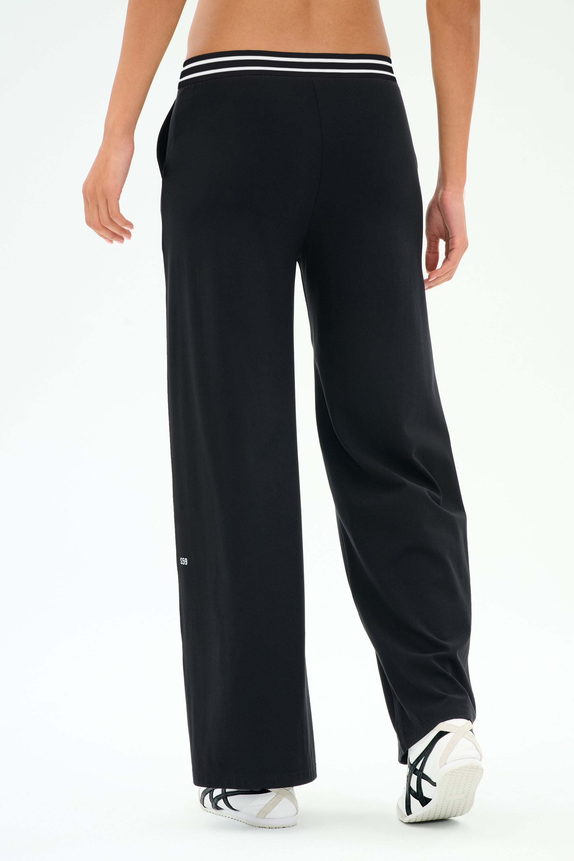 Someone stands facing away, wearing SPLITS59 Michele Airweight Trouser in black with a white-striped waistband and white sneakers against a plain backdrop.