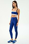A woman stands against a plain background wearing the SPLITS59 Monah Rigor Bra in Cobalt/Black, with matching blue high-waisted leggings featuring black stripes, and blue athletic shoes.