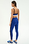 A woman faces away against a white background, wearing the SPLITS59 Monah Rigor Bra in Cobalt/Black with matching leggings featuring black side stripes, and blue Adidas sneakers.