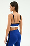 A woman stands facing away, showcasing the back of her SPLITS59 Monah Rigor Bra - Cobalt/Black and matching leggings against a plain background.